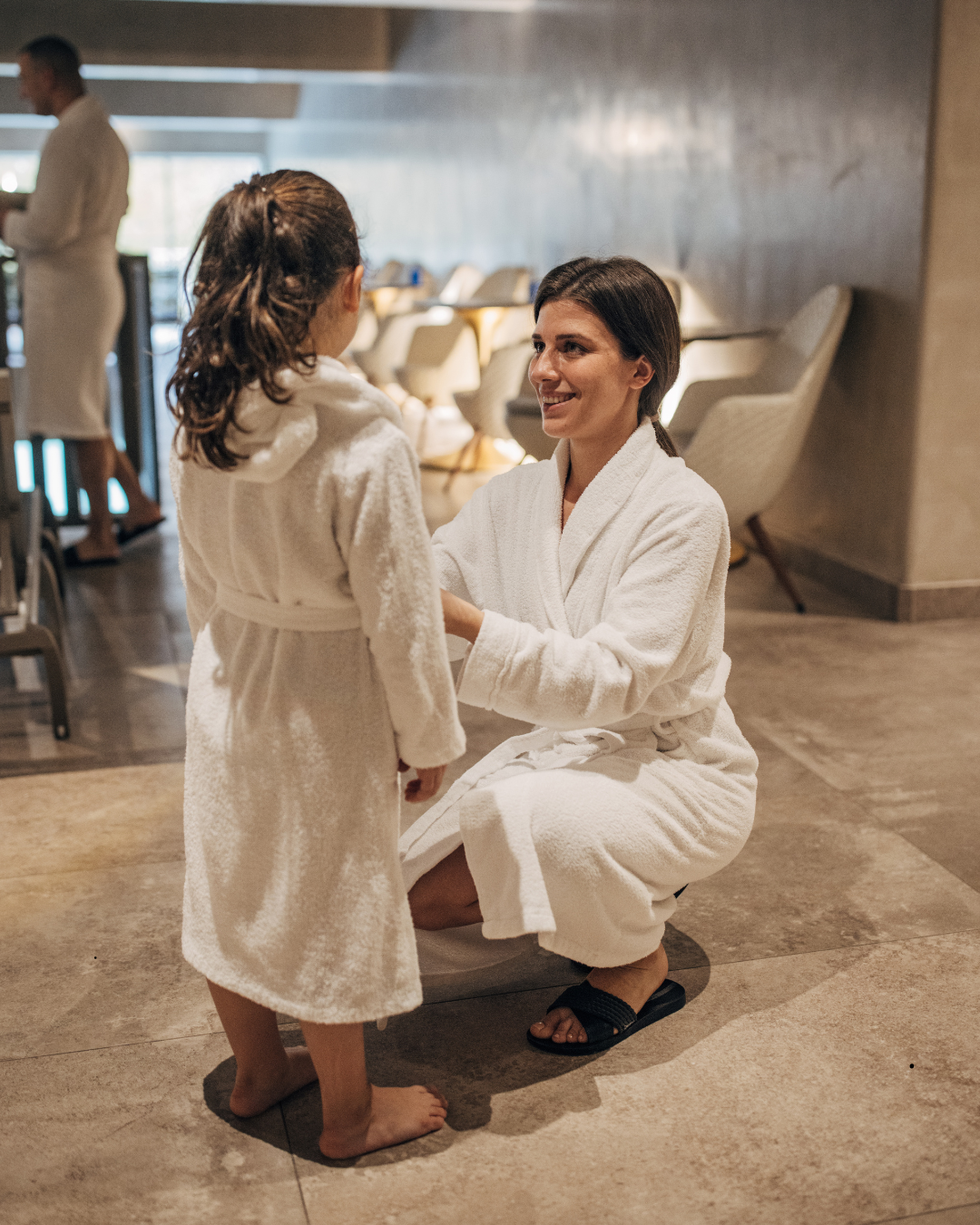 Une femme en peignoir blanc tend la main à une jeune fille en peignoir blanc dans un spa ou sauna, avec un mobilier confortable en arrière-plan.