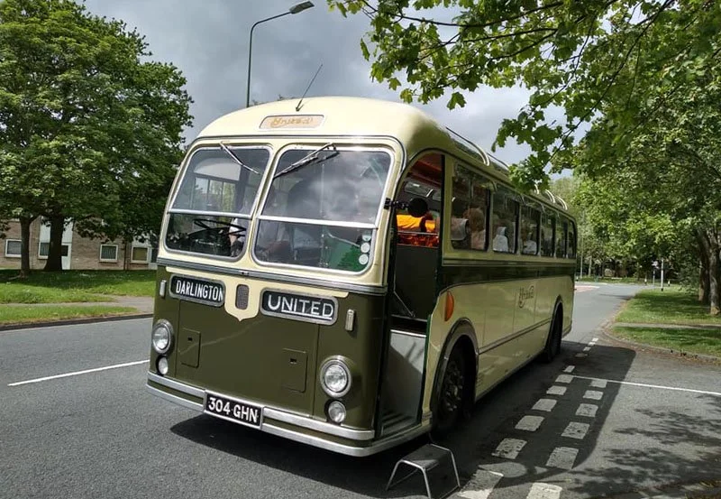 Vintage bus painted in green and cream, parked on a suburban street with trees and houses in the background. The bus has signs indicating "Darlington" and "United" and a license plate reading "304 GHN."