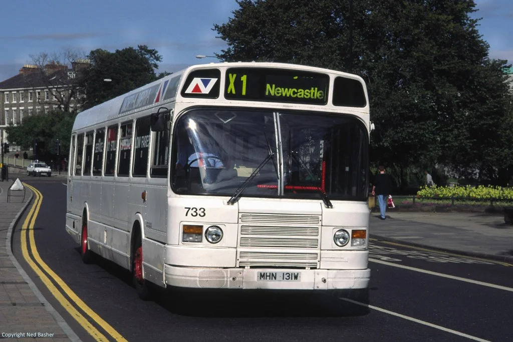 A white bus on a city street with a sign indicating route X1 to Newcastle, with trees and buildings in the background.