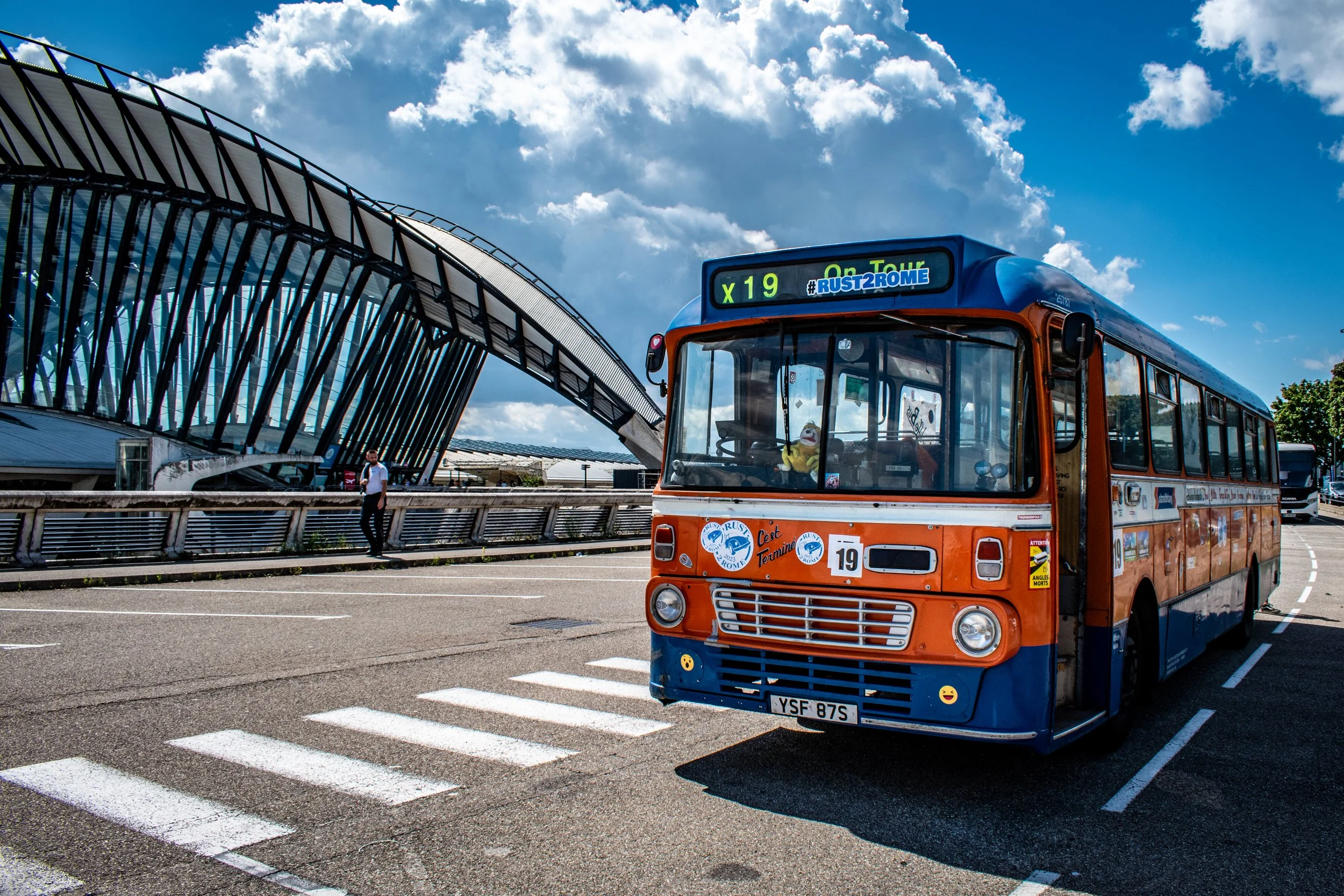 A vintage orange and blue bus with a puppet inside, parked at a curb near a modern architectural building, under a partly cloudy sky.
