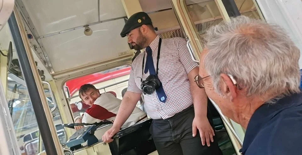 Three men inside a vintage bus. The man in the driver's seat, wearing a beige jacket with maroon stripes, is looking back and smiling. The other two men are standing and talking; one with a camera hanging around his neck, and the other with gray hair and glasses.