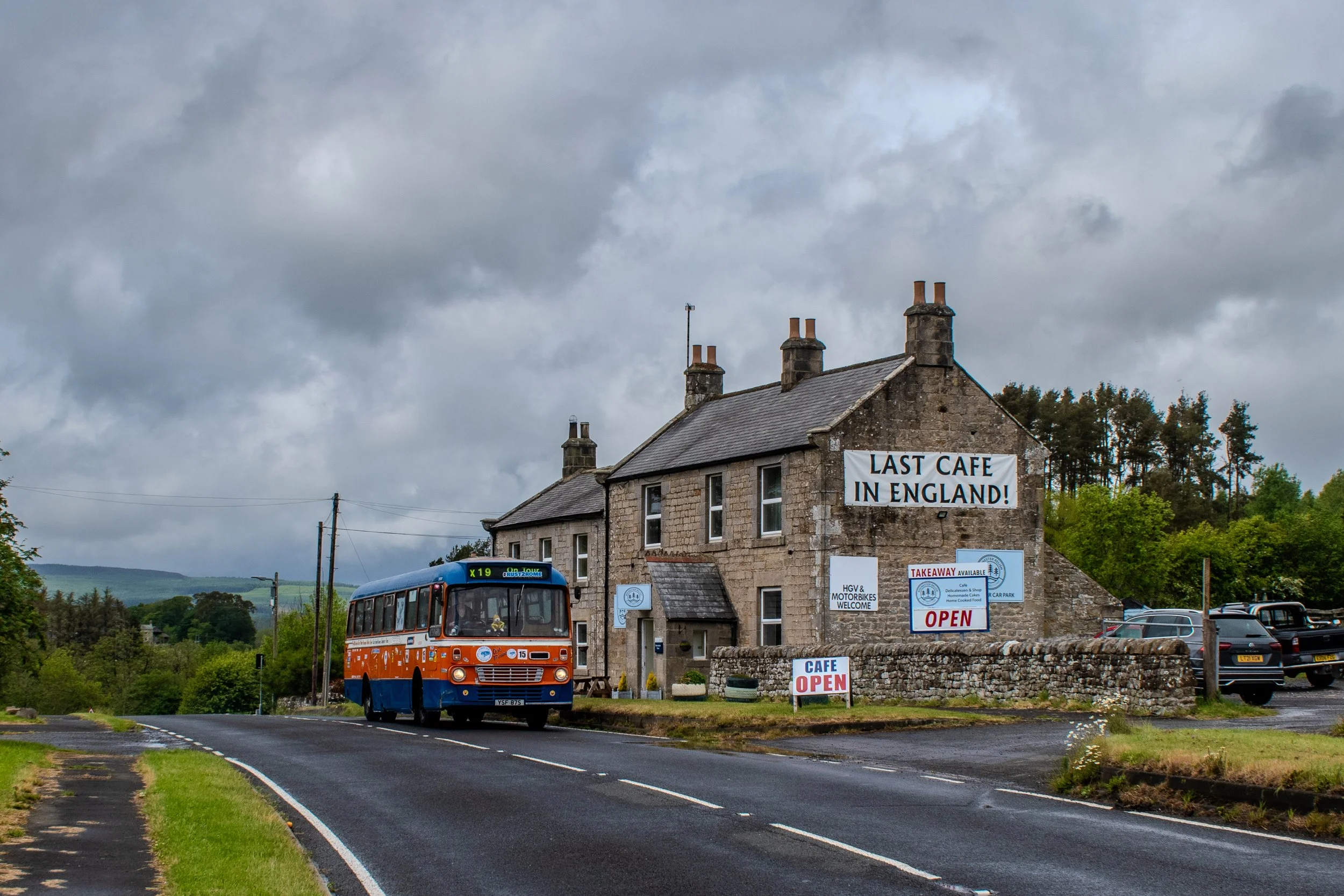 A vintage blue and orange bus driving past a rustic stone building with signs advertising a cafe, which is open and the last cafe in England. The building is on a rural road under a cloudy sky surrounded by greenery and trees.