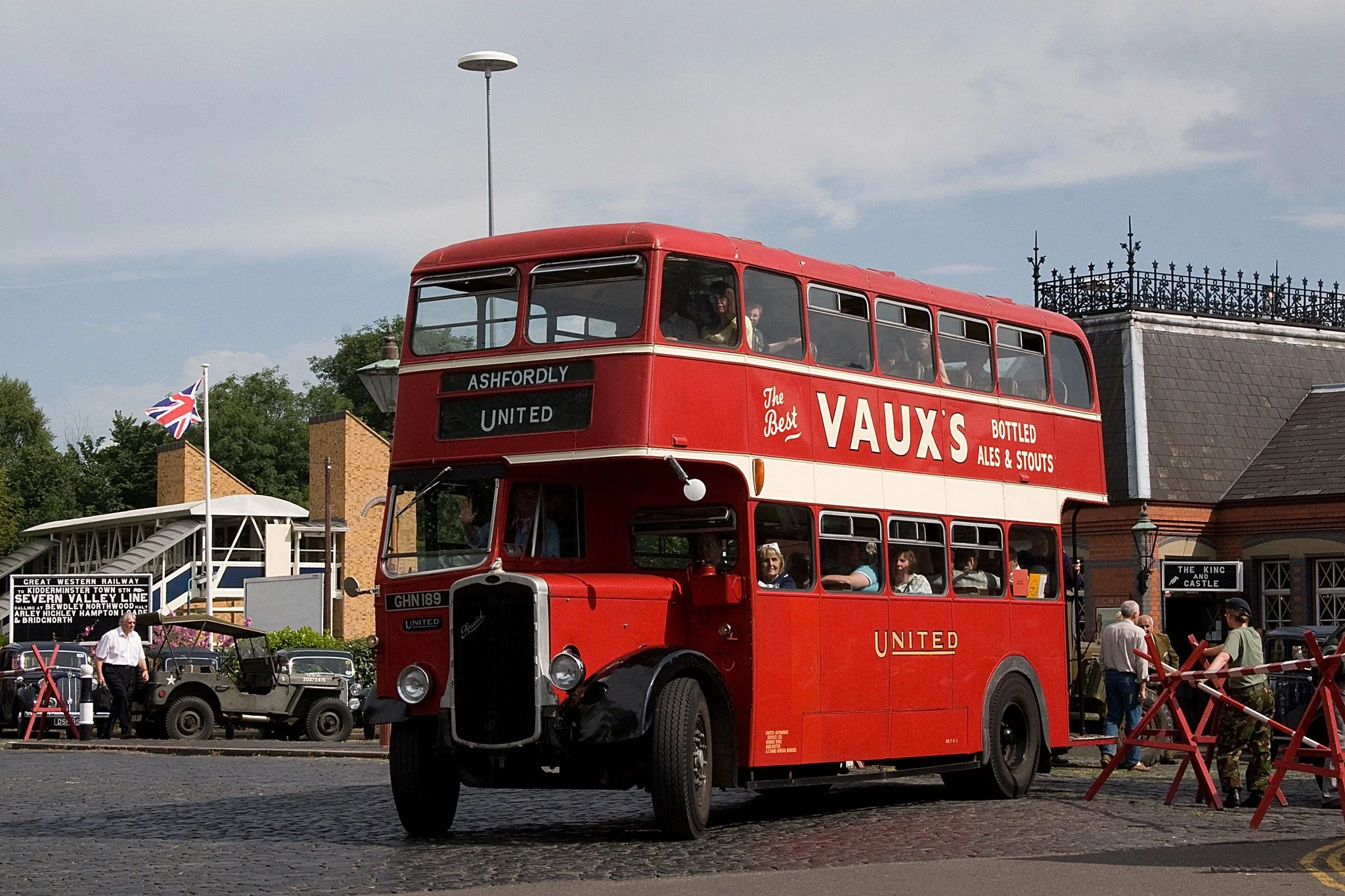 A vintage red double-decker bus with advertisements for Vaux's ales and stouts, parked outdoors during daytime. People are visible inside and nearby, with a historic-looking building and a British flag in the background.