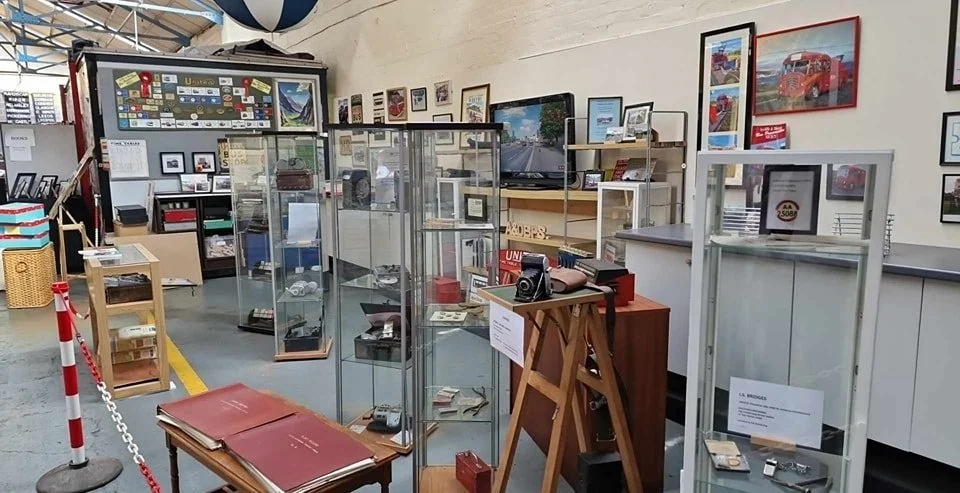 An indoor museum exhibit featuring glass display cases with vintage items like cameras, phones, and artifacts, surrounded by framed pictures on the wall and a television