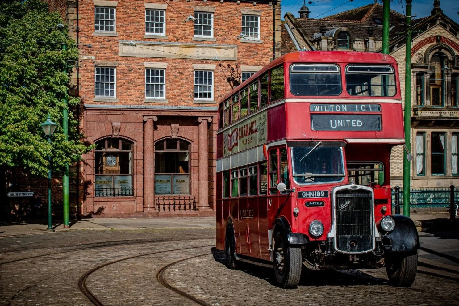 An old red double-decker bus on a street, with trees and a park in the background, and cloudy sky.