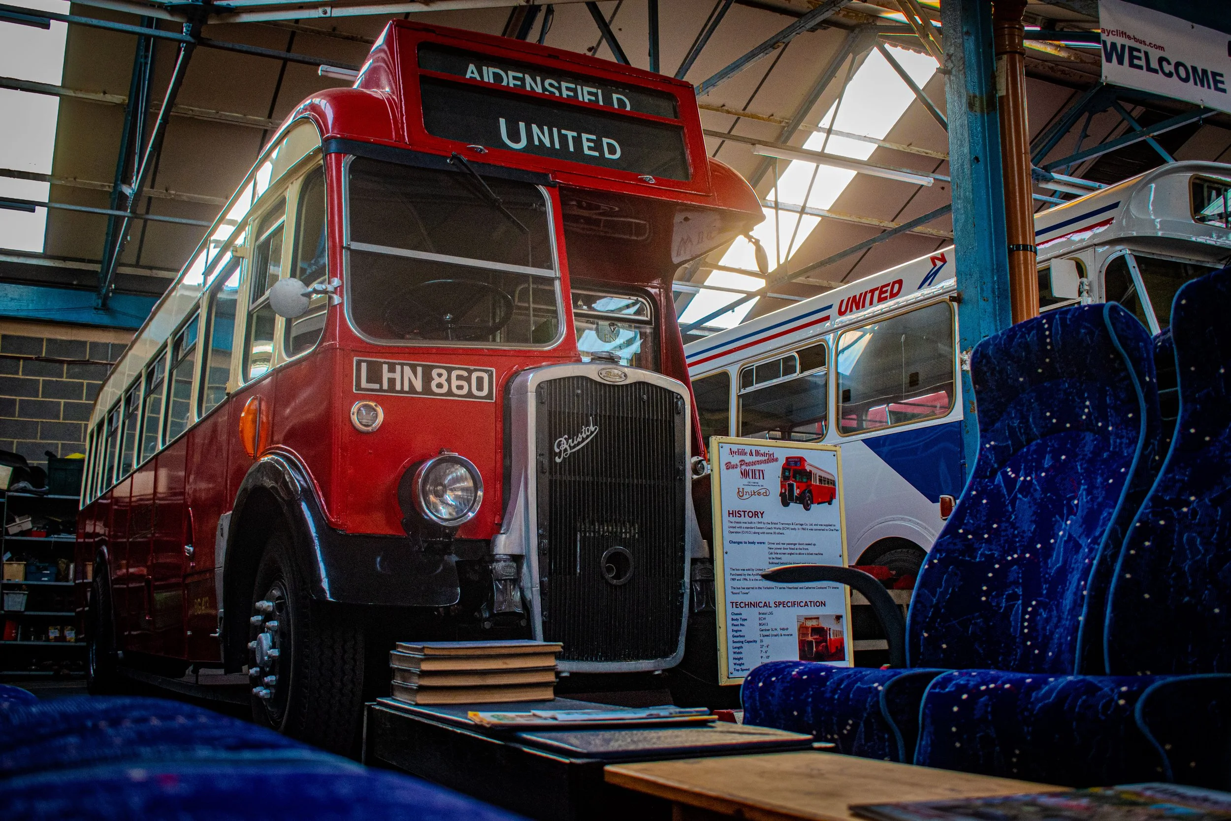 Vintage red and white bus with a sign displaying 'AIDENSFIELD UNITED,' on display at an indoor museum or exhibit, with informational placards in front and seats nearby.