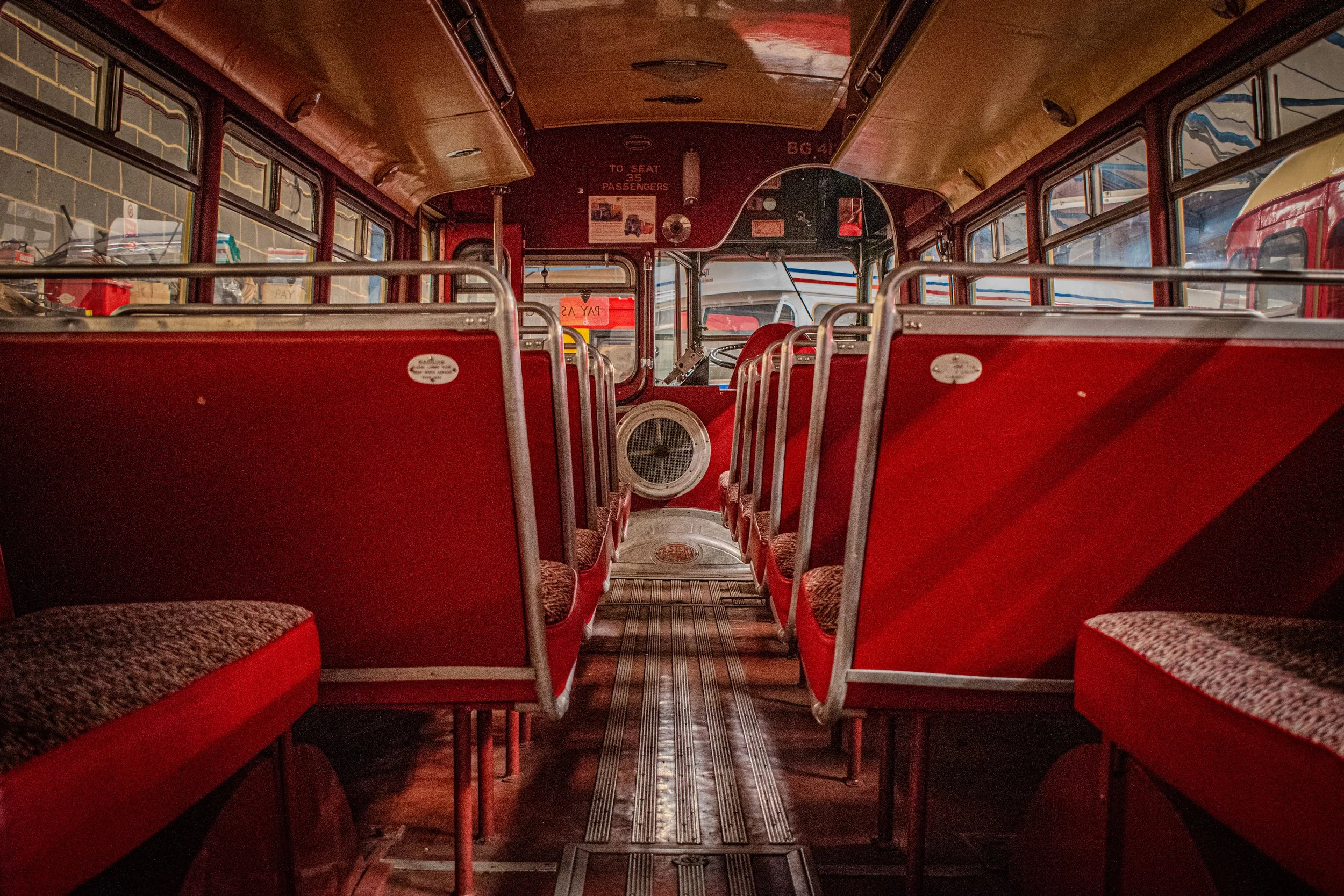 Interior of a vintage red bus with red seats, brown patterned cushions, metal handrails, a speaker near the front, and a driver’s area visible through the windshield.