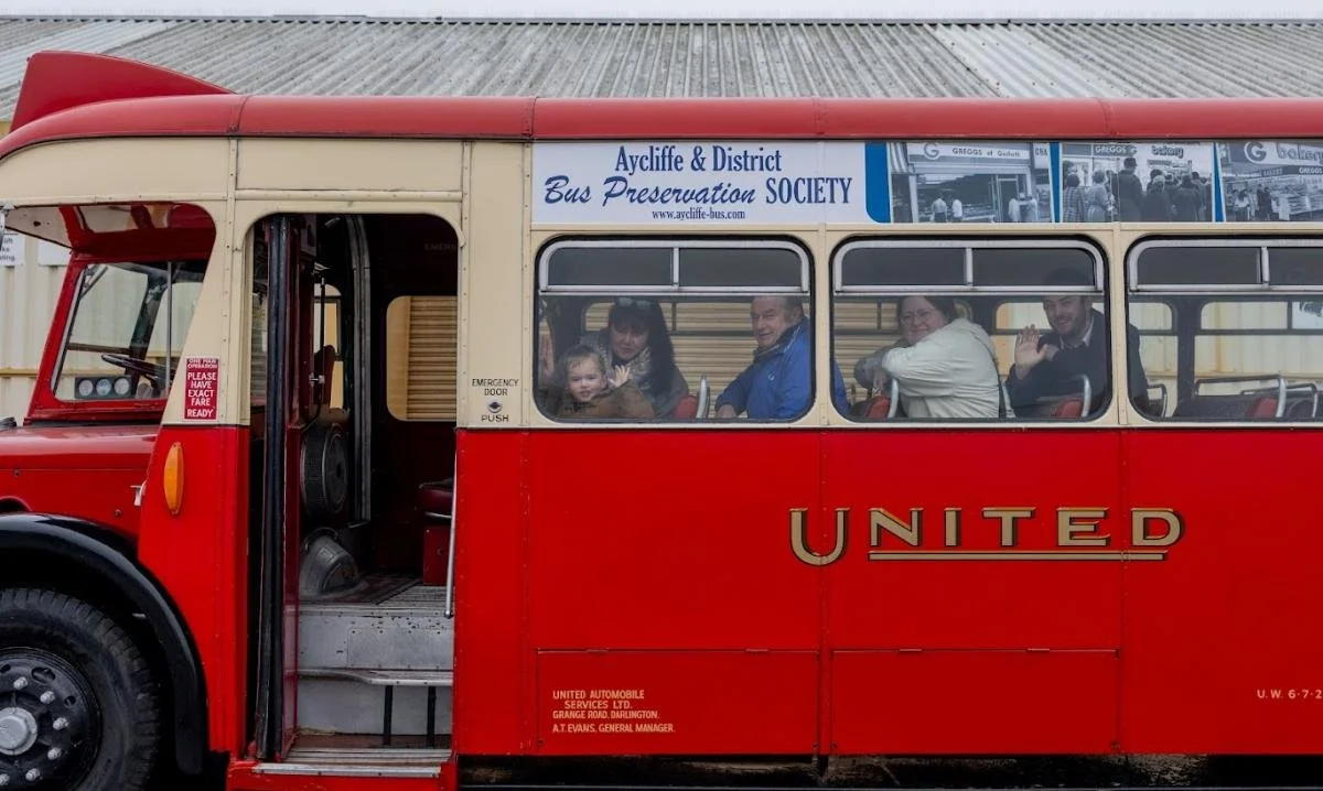 A vintage red and cream bus with passengers inside, including a child and adults, parked outdoors. A sign on top reads 'Aycliffe & District Bus Preservation Society.'