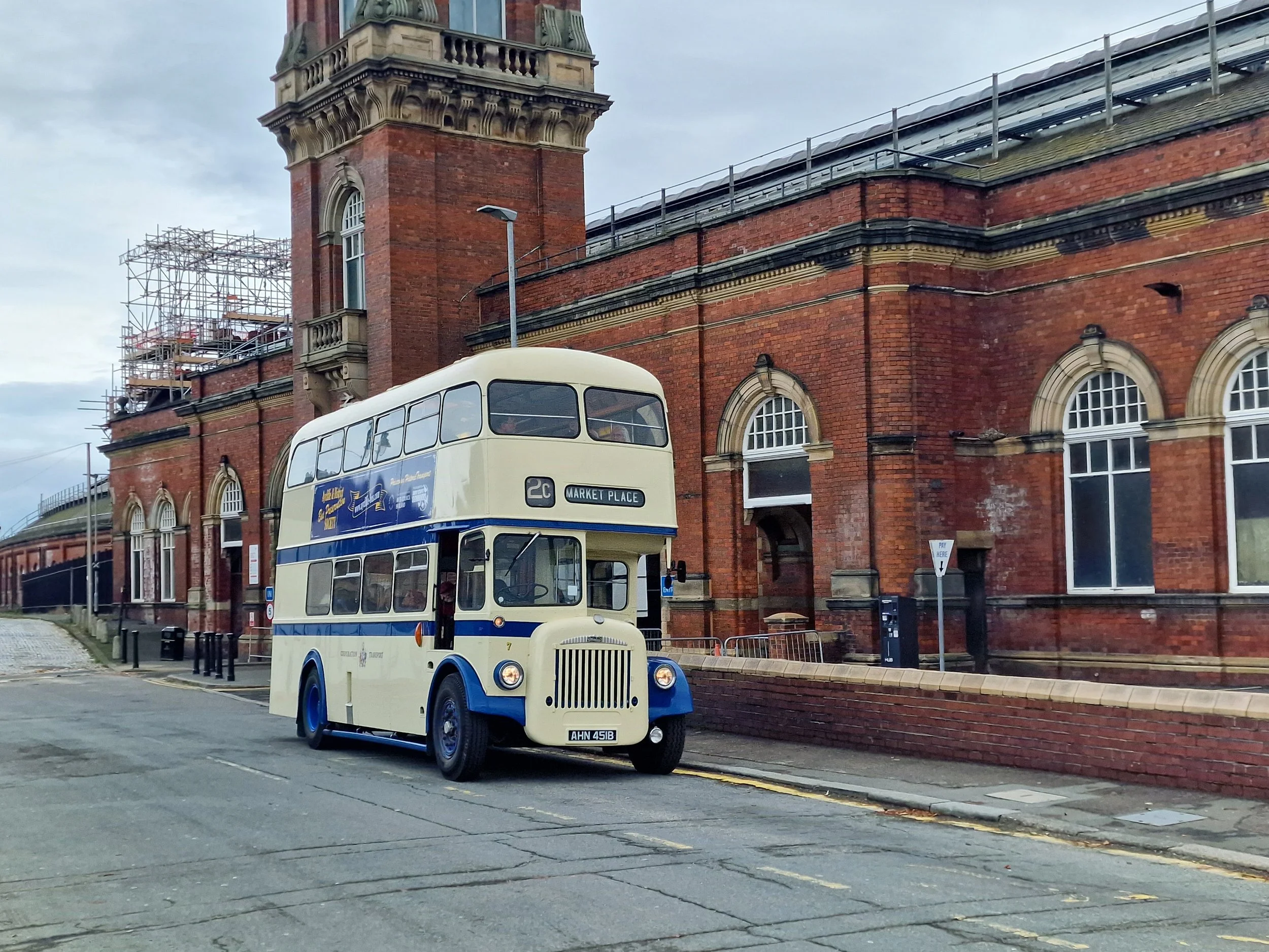 A vintage double-decker bus parked on a street in front of a red brick building with arched windows and a clock tower.