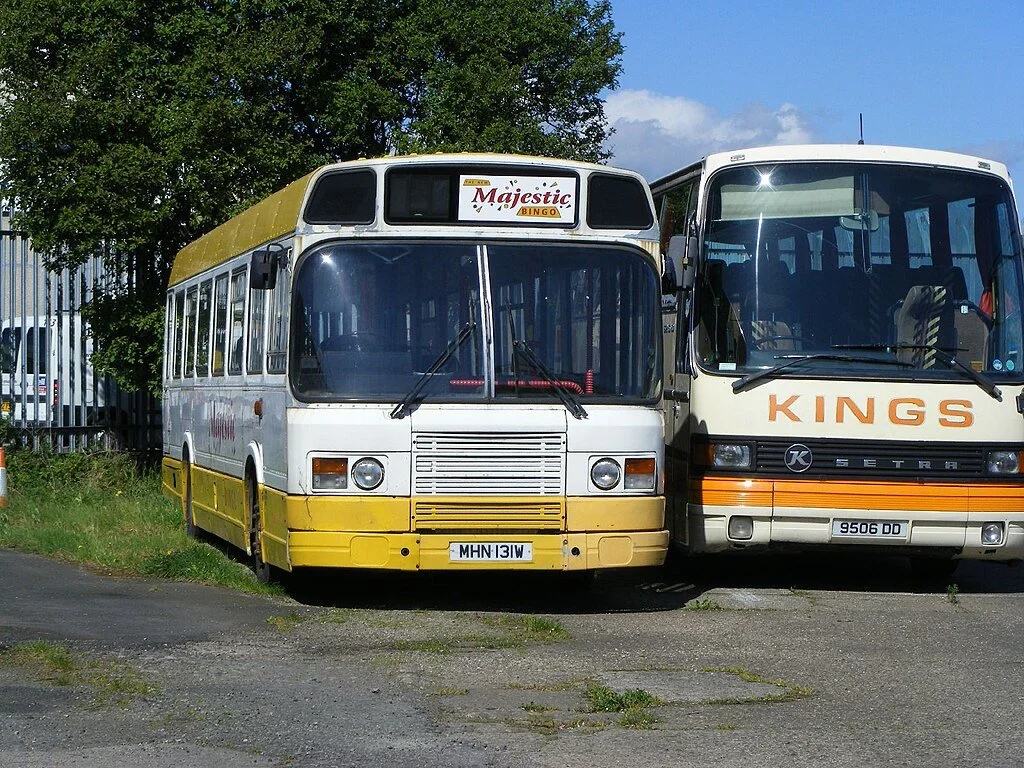 Two buses parked side by side, one older with a yellow and white color scheme and a sign for 'Majestic Bingo' on the front, and another newer white bus with 'KINGS' written on the front.