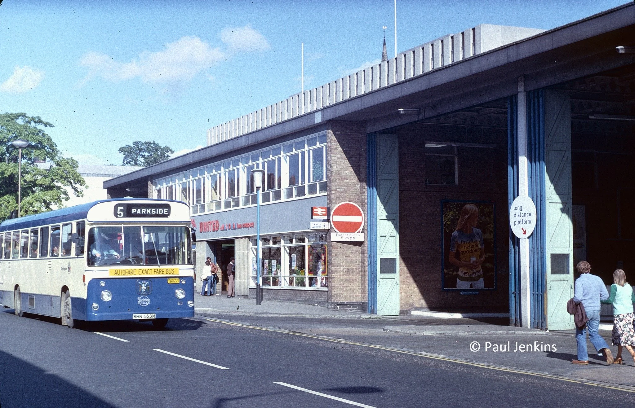 A vintage bus arriving at a bus stop in an urban area, with a brick building, several pedestrians, and street signs visible.