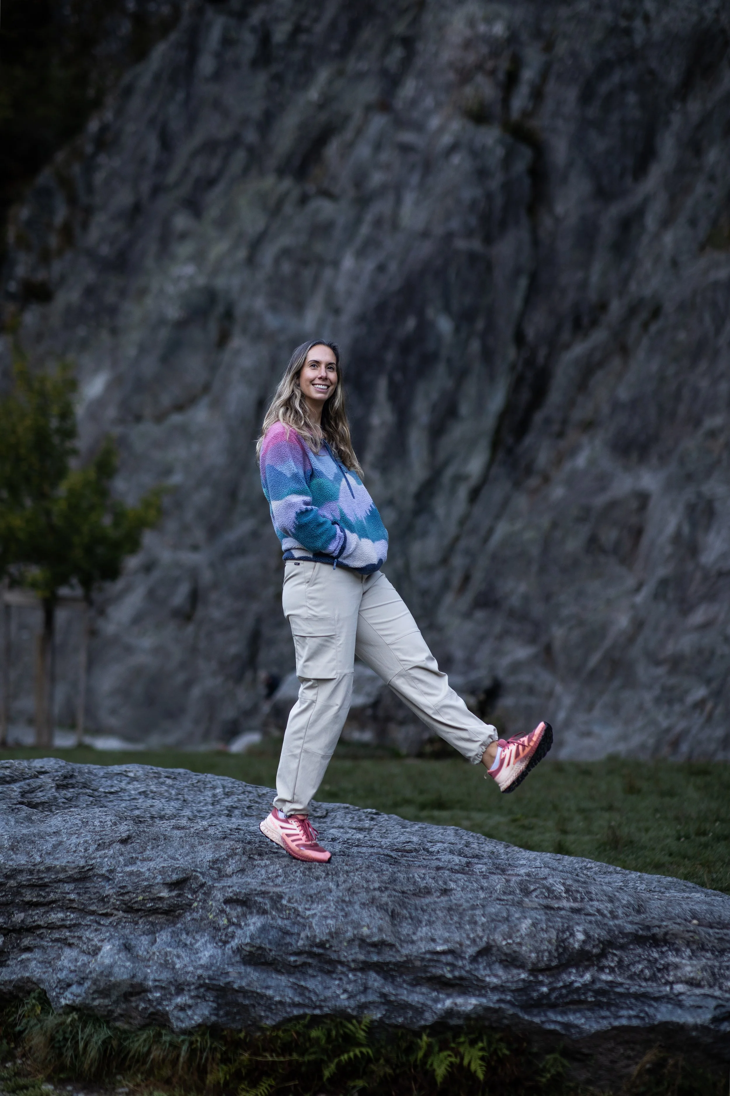 image of woman standing on a rock with one leg kicking forward wearing a fleece and pink trainers and combat trousers smiling and looking at the camera