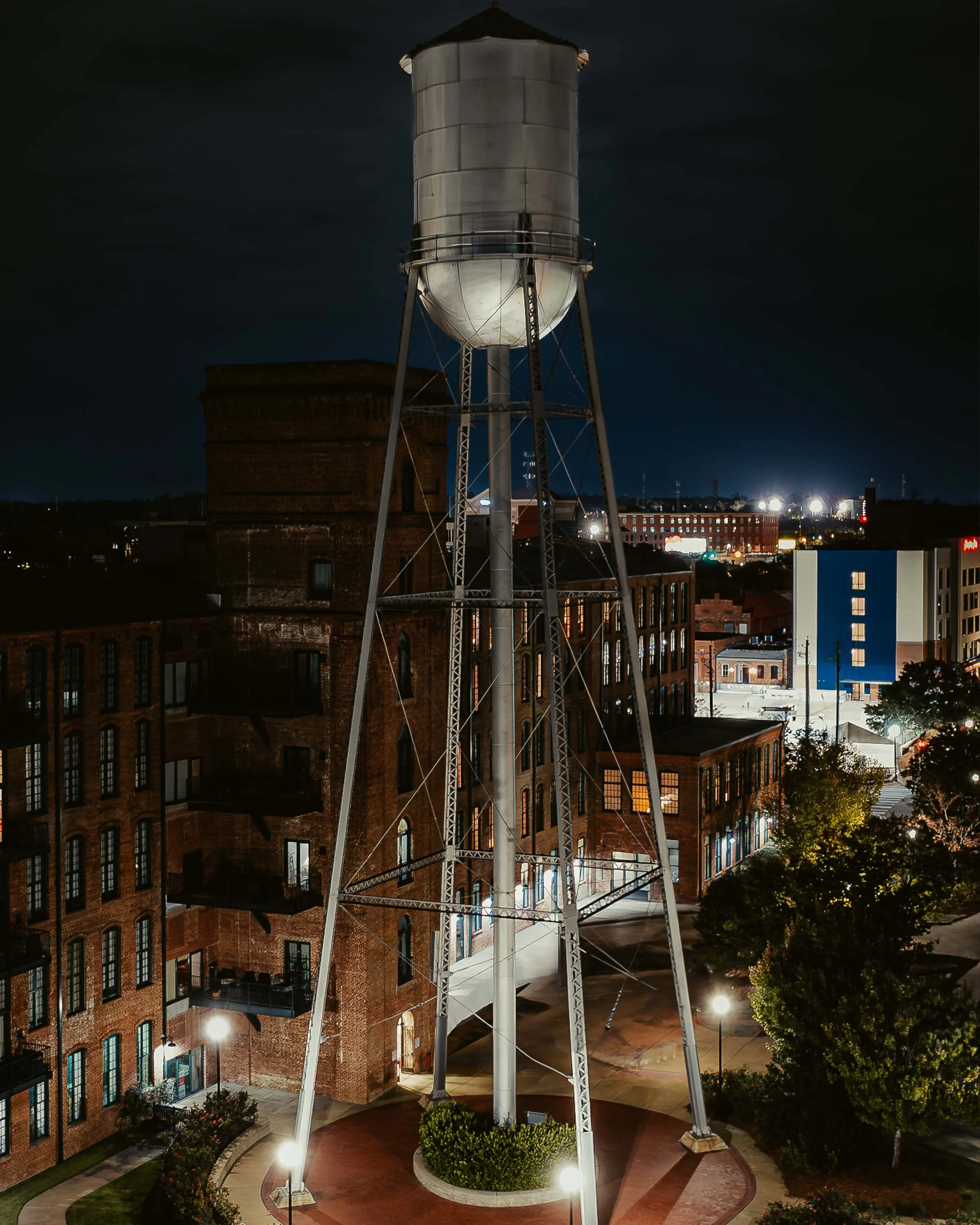 Night view of a water tower in an urban area with illuminated streets, brick buildings, trees, and city lights in the background.