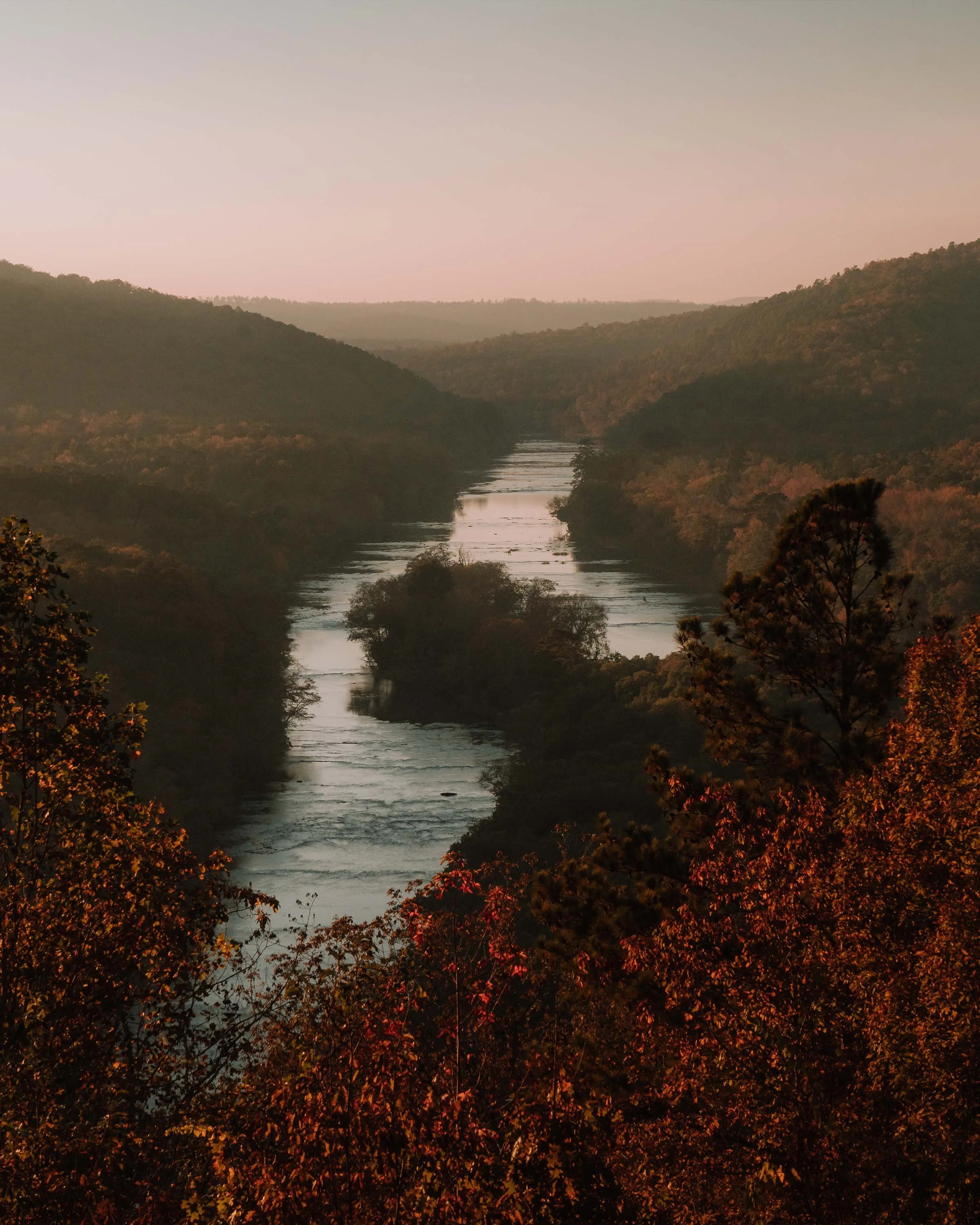 A river flowing through a lush, forested valley during sunset, with hills on either side and colorful trees in the foreground.