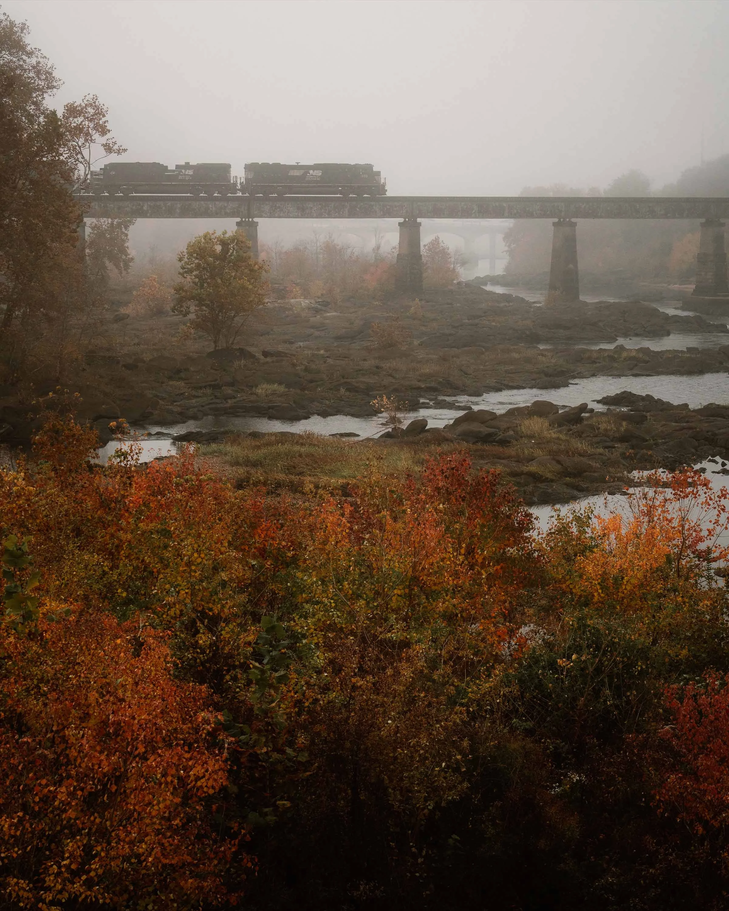 A train traveling on a bridge over a rocky riverbed during foggy autumn day, surrounded by colorful fall foliage.