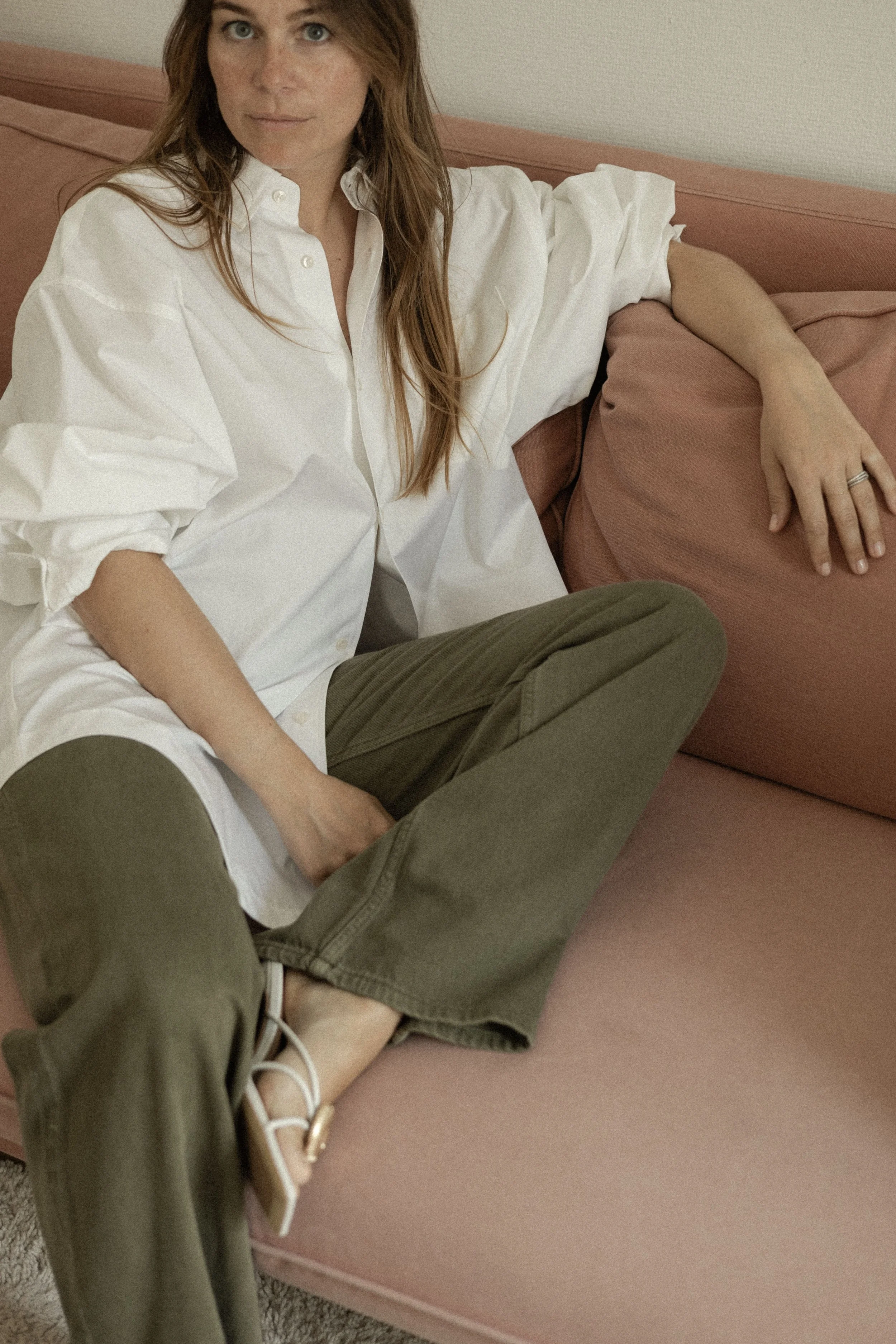 Alexandra BAYLE - Femme aux cheveux longs, habillée d'une chemise blanche, assise sur un canapé rose, portant des pantalons kaki, portant des sandales blanches.