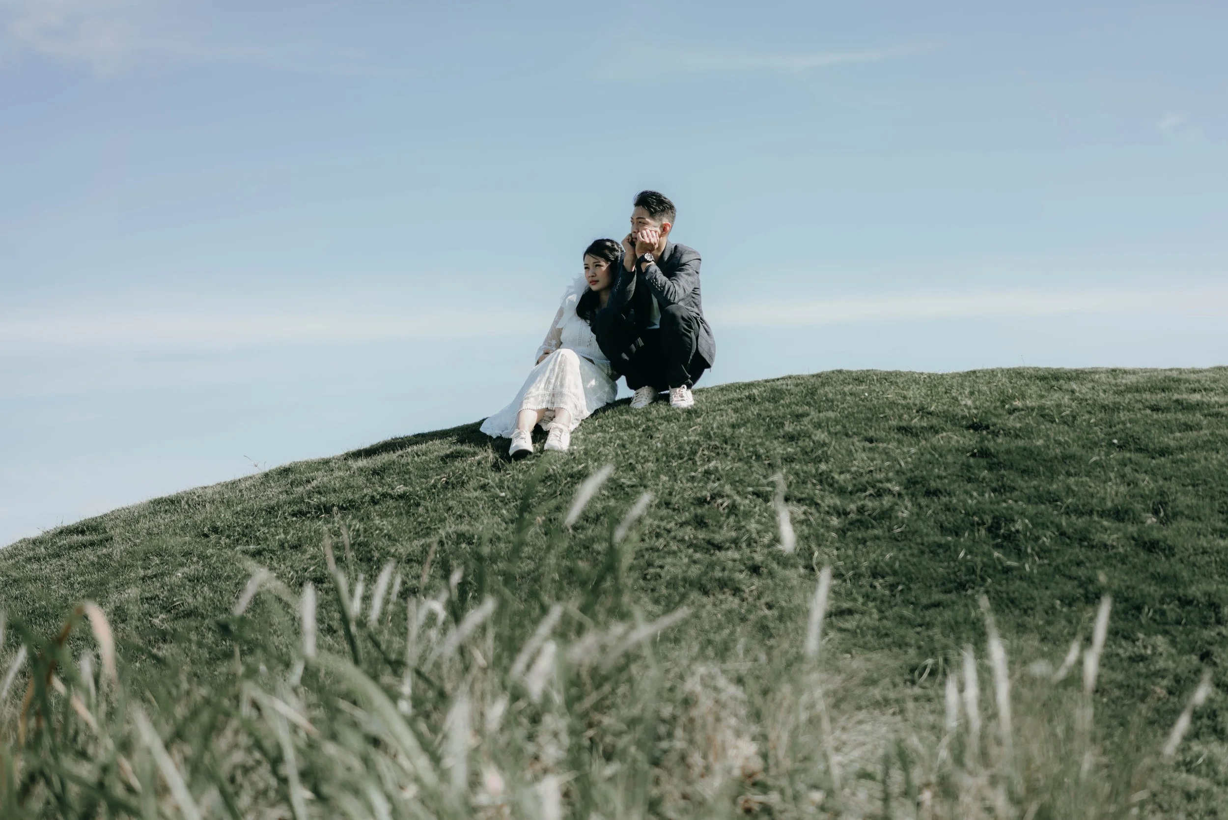 A couple sitting on a grassy hilltop under a blue sky, with the woman dressed in a white dress and the man in dark clothing.