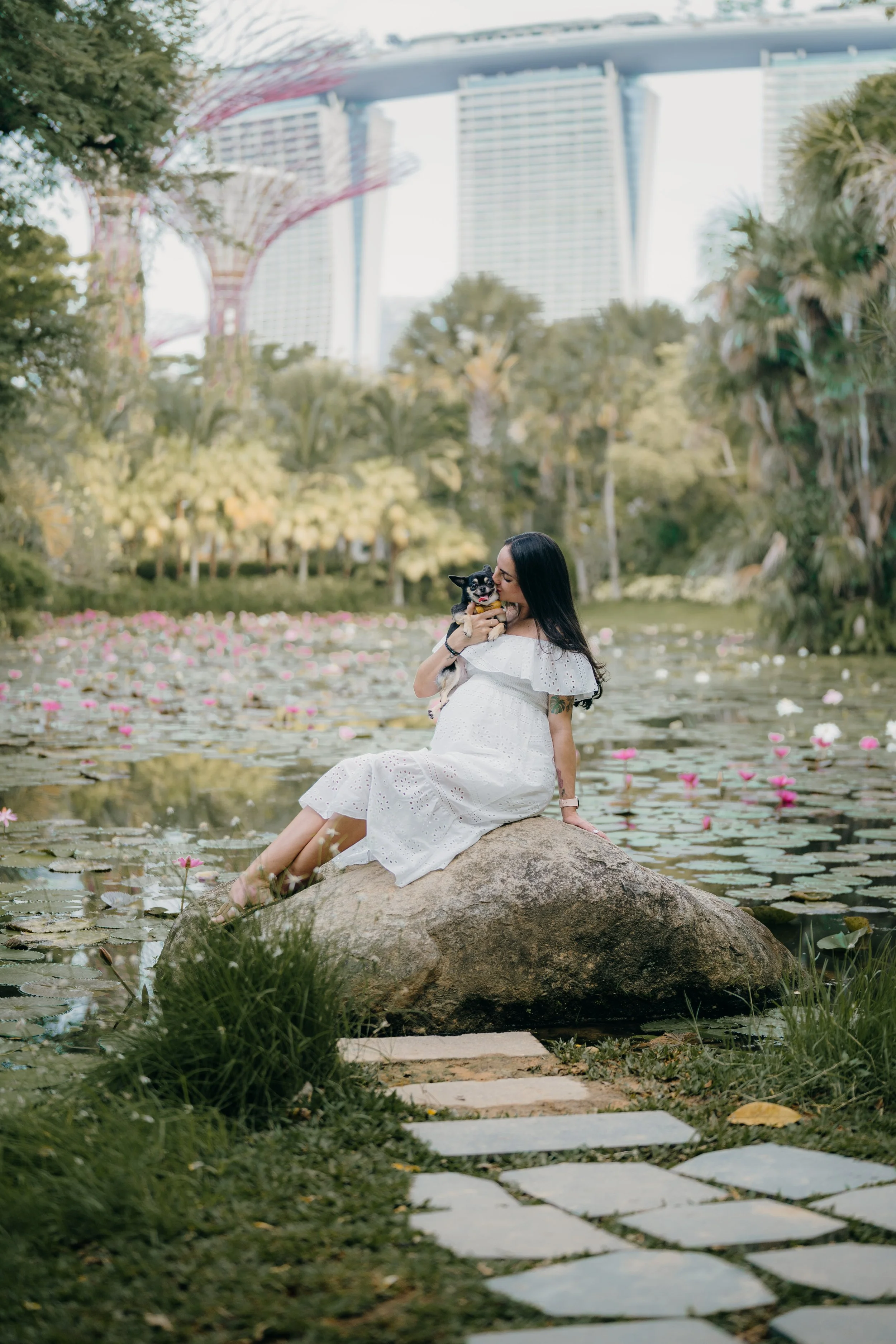 A woman in a white dress sits on a large rock in a pond with pink and white water lilies, holding a small dog in her arms. The background features lush greenery and tall buildings, including the Marina Bay Sands in Singapore.