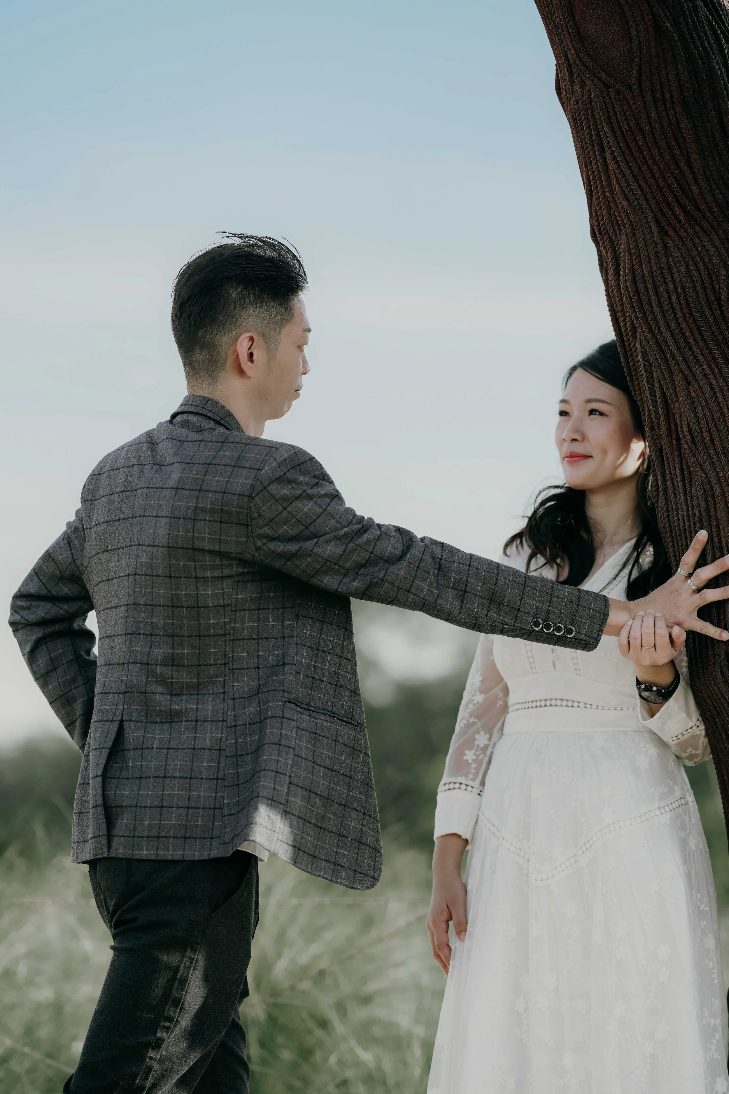 A man in a gray checked blazer touching a woman in a white dress, outdoors near a tree with a cloudy sky in the background.