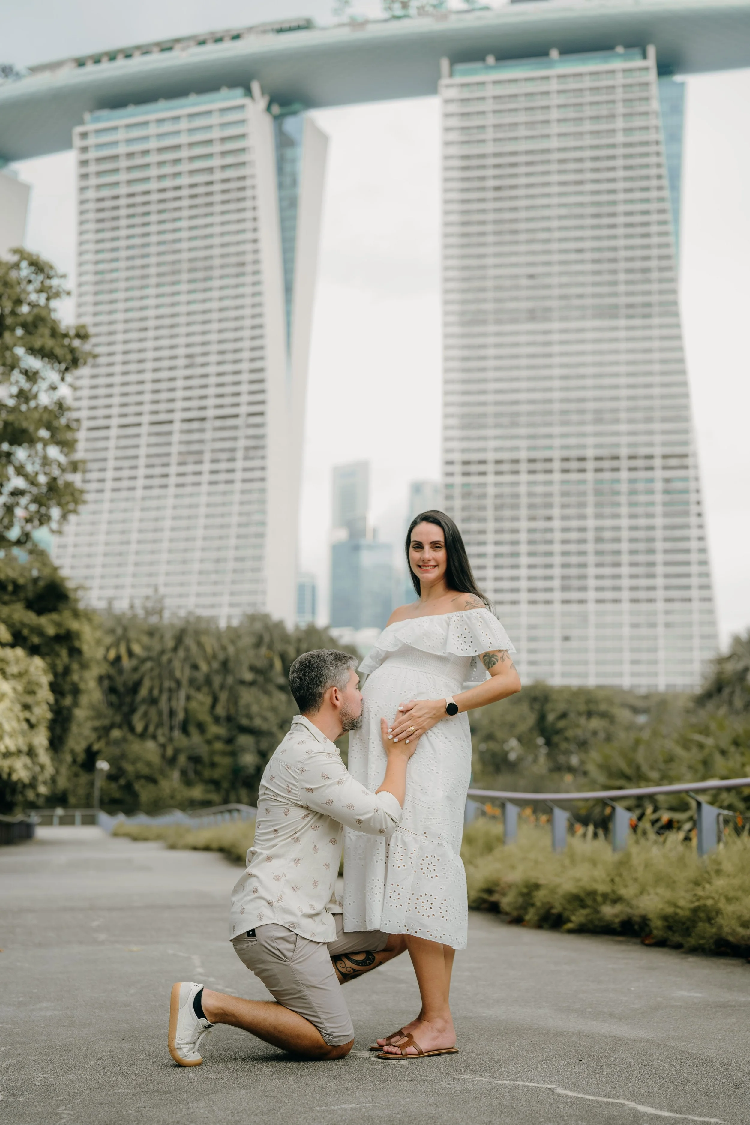 A man proposes to a pregnant woman outdoors with the Marina Bay Sands hotel in Singapore in the background. The woman is wearing a white dress and smiling, while the man is kneeling, holding her belly, and looking up at her.