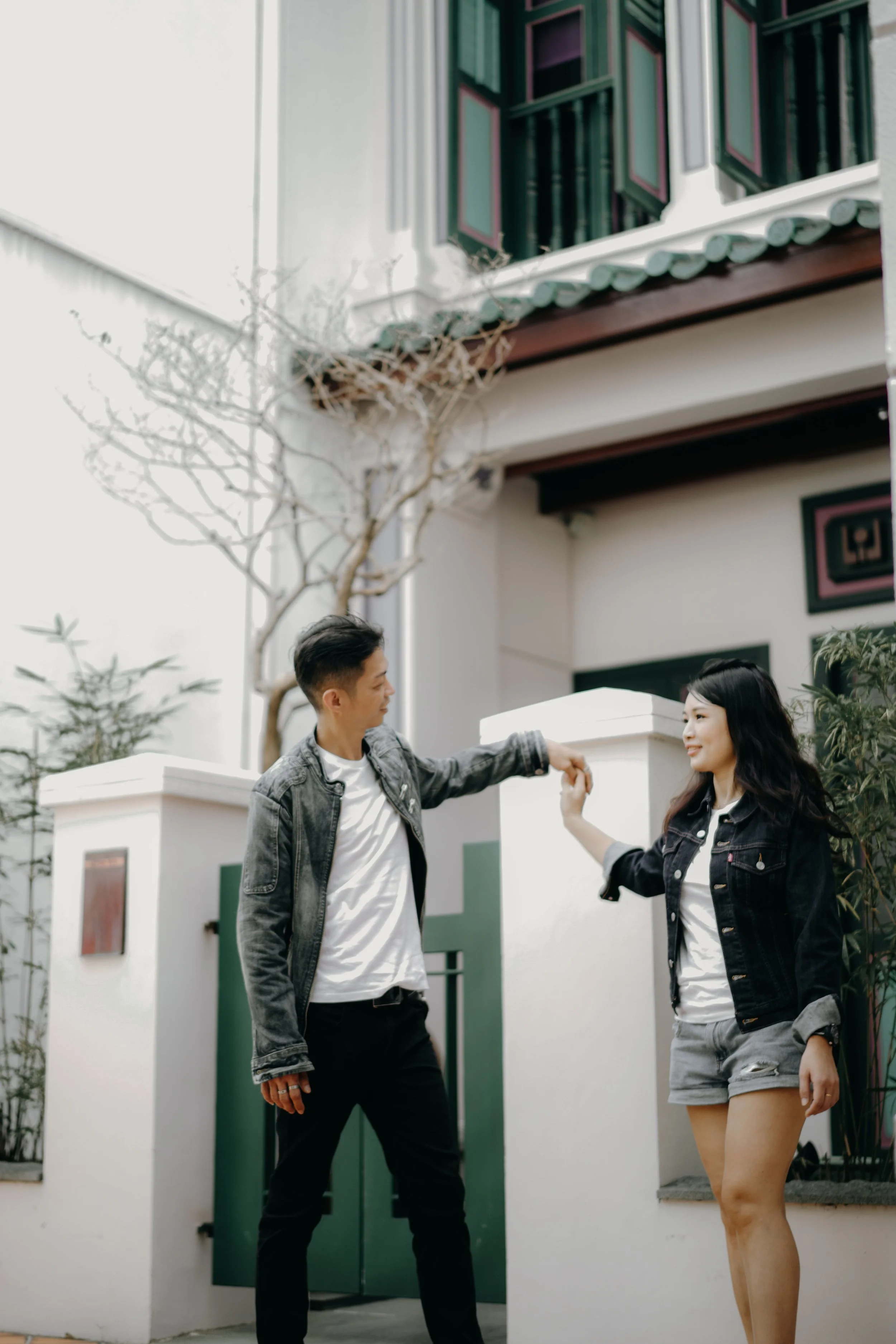 A young man and woman dance outside in front of a white house with a green door, holding hands and smiling.