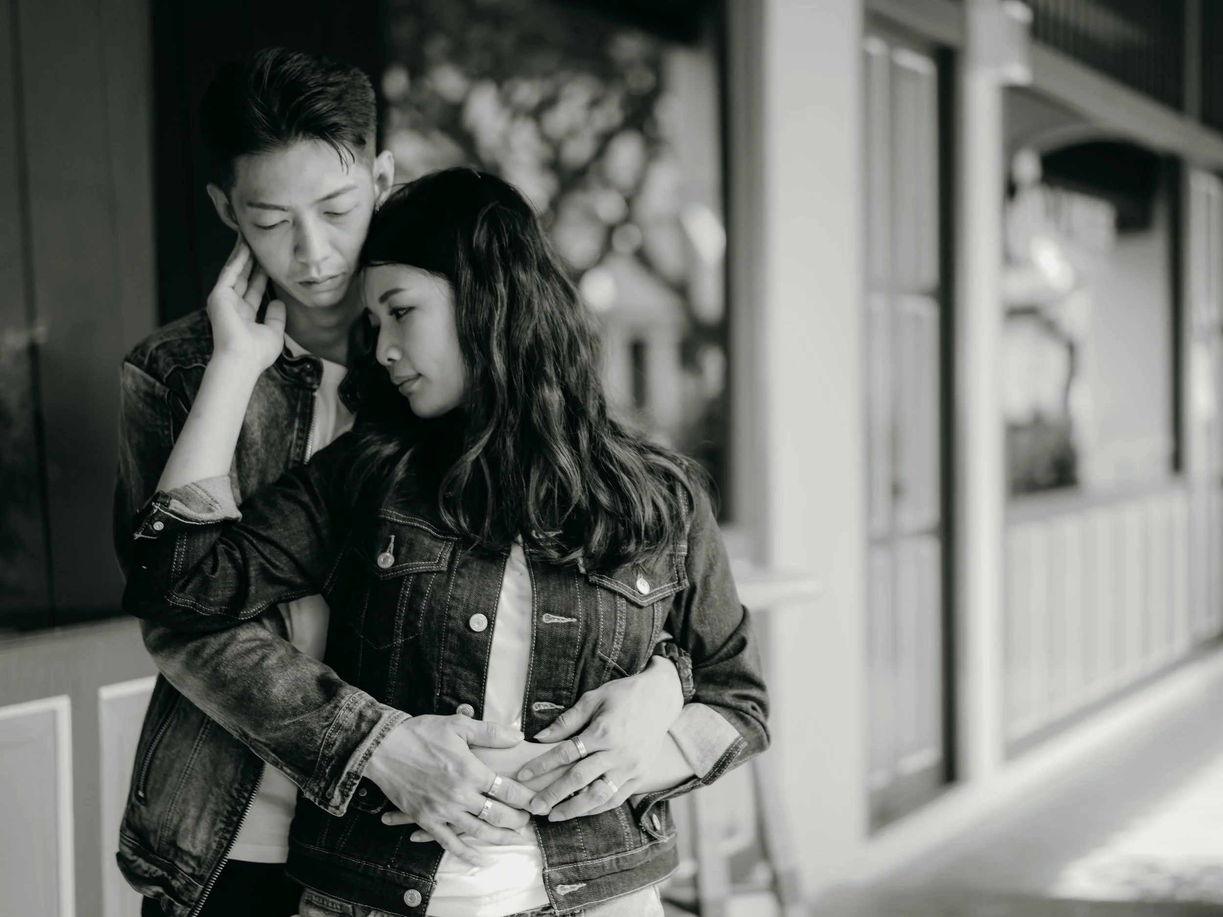 A young man and woman in an embrace with their foreheads touching, standing outdoors near a building with large windows, in black and white.