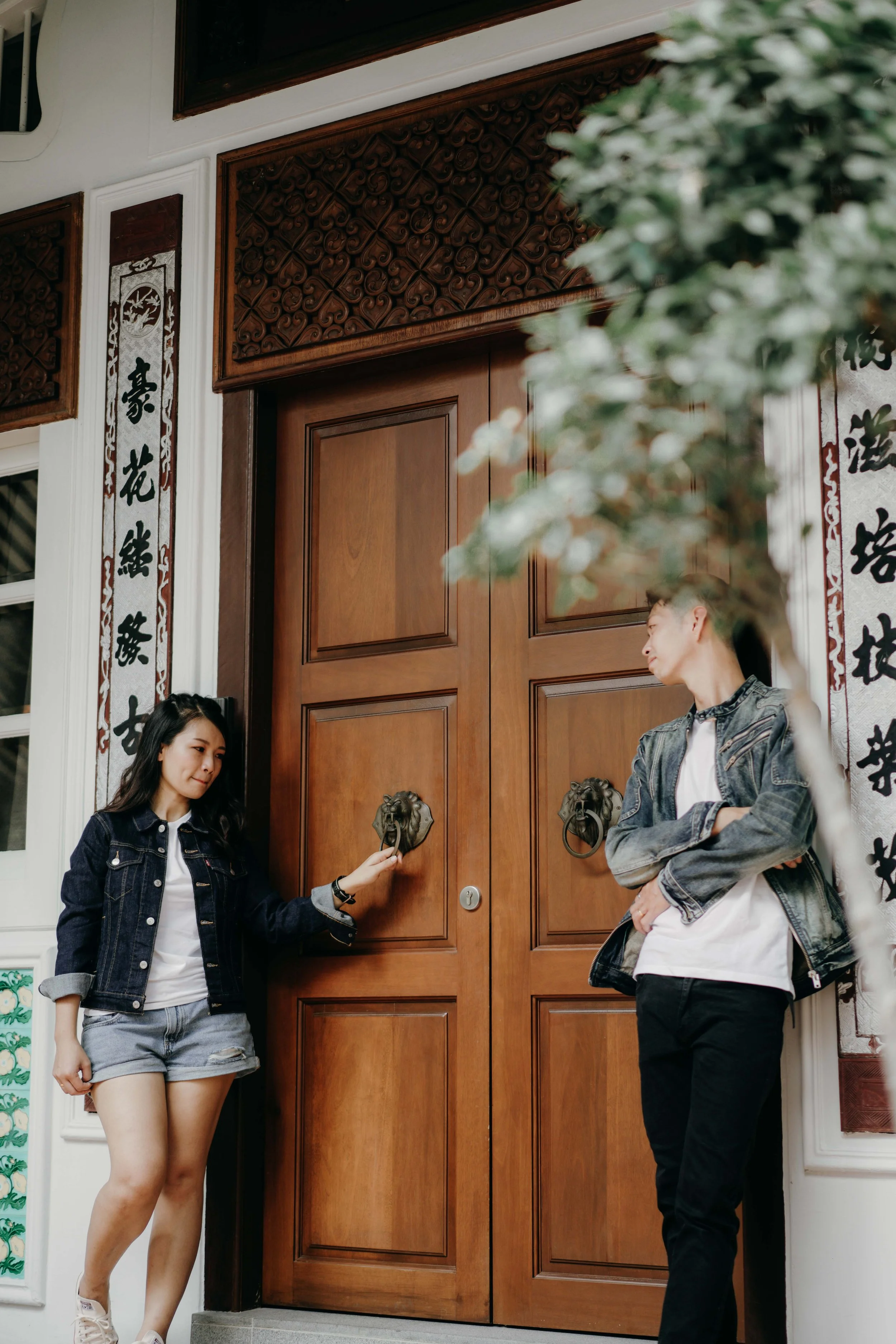 A young woman and a young man standing in front of a large wooden door with lion head knockers. The woman is holding a door knocker and the man is leaning against the wall with arms crossed. Both are casually dressed, the woman in a denim jacket and 