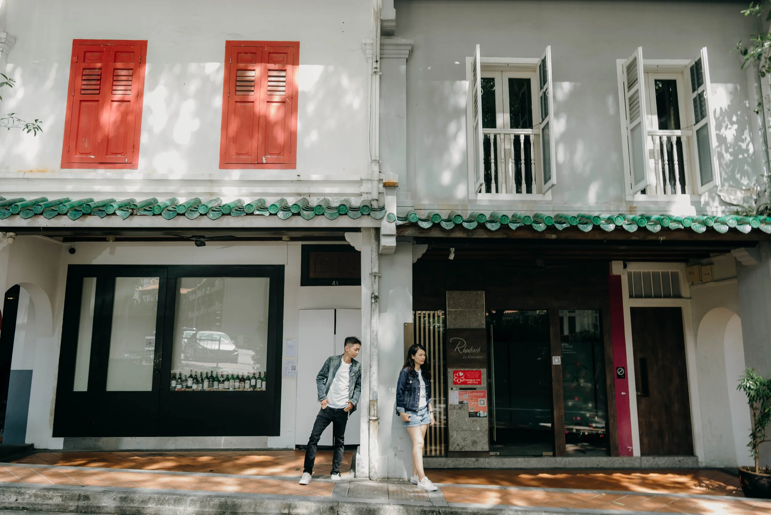 Two young people standing on a sidewalk in front of a white building with red shutters and open white window shutters, with shadows of tree leaves on the facade.