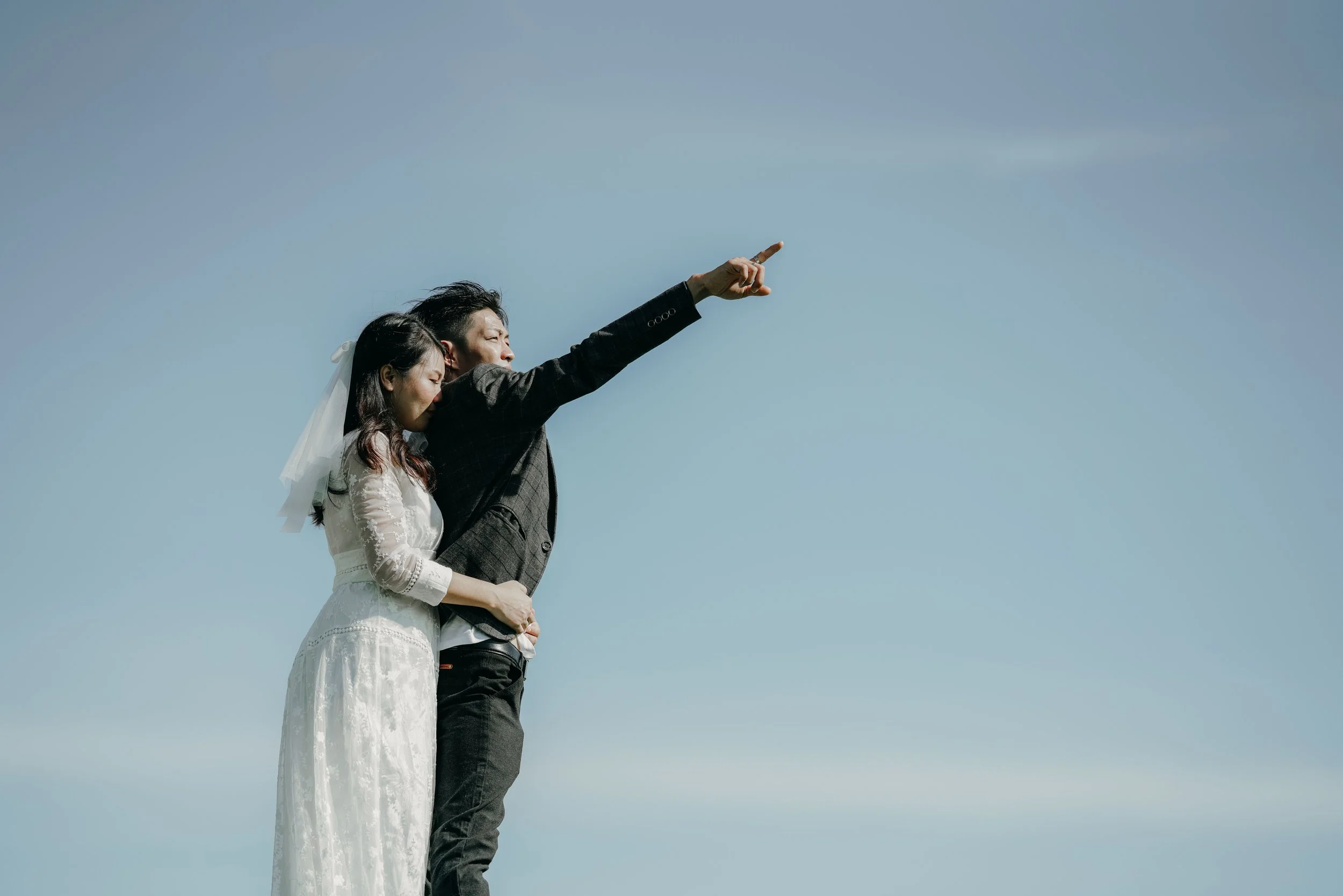 A man in a suit and a woman in a white dress stand against a clear blue sky. The man is pointing into the distance, and the woman is leaning on his shoulder with her eyes closed.