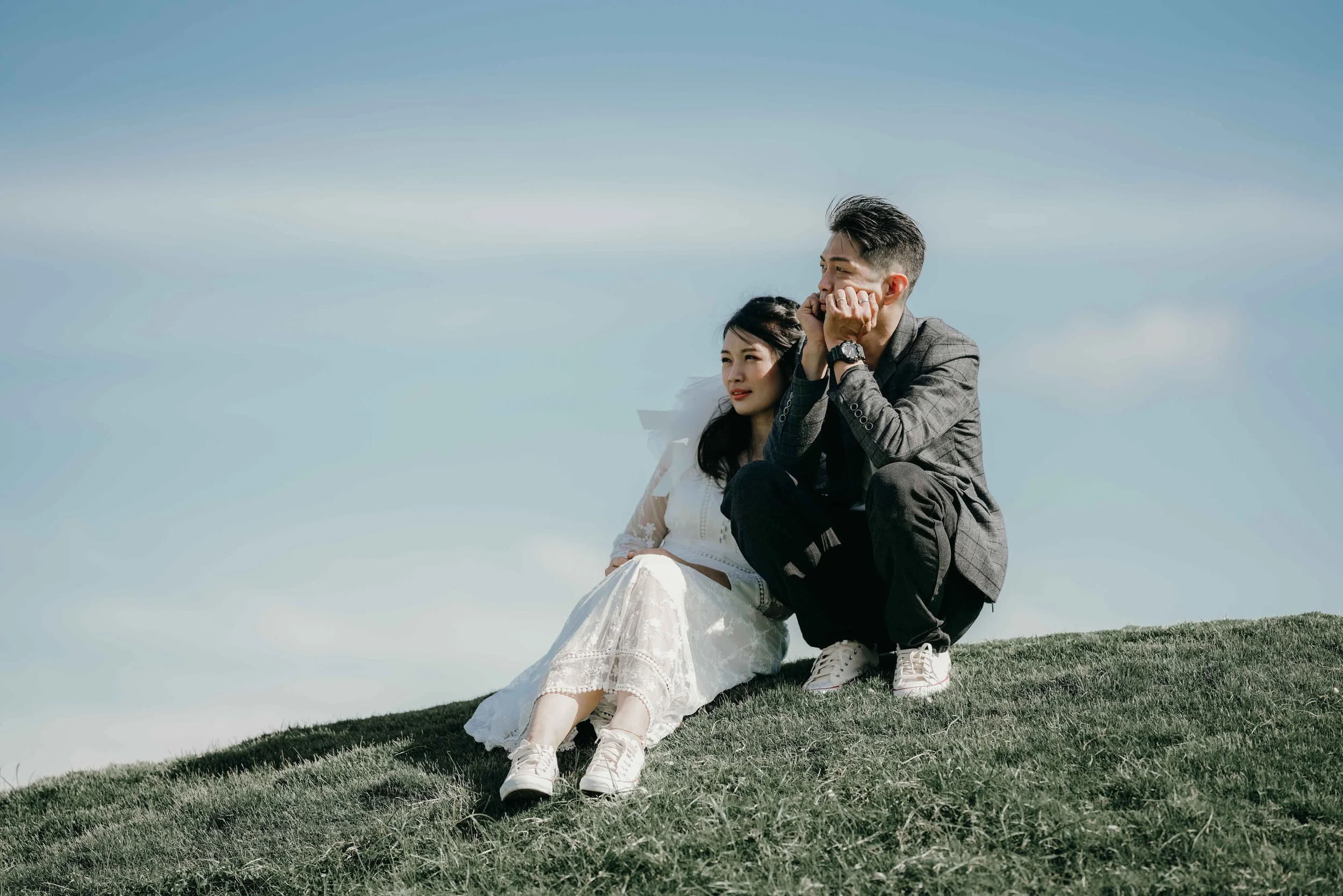 A young couple sitting on a grassy hilltop under a clear blue sky, with the woman in a white dress and the man in a gray blazer, both wearing sneakers, enjoying a peaceful moment together.