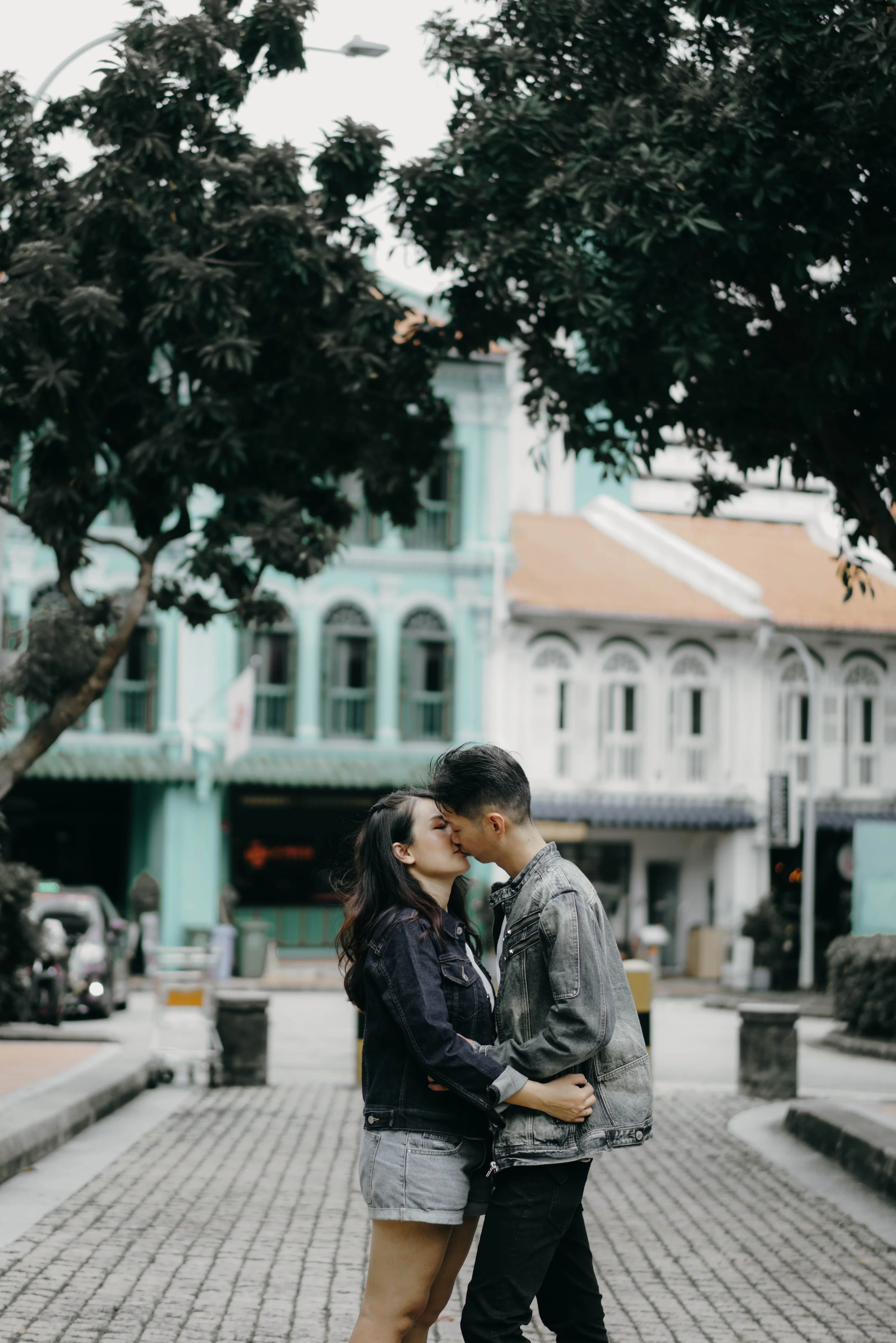 A young couple kissing on a street with vintage-style colorful buildings and large trees in the background.
