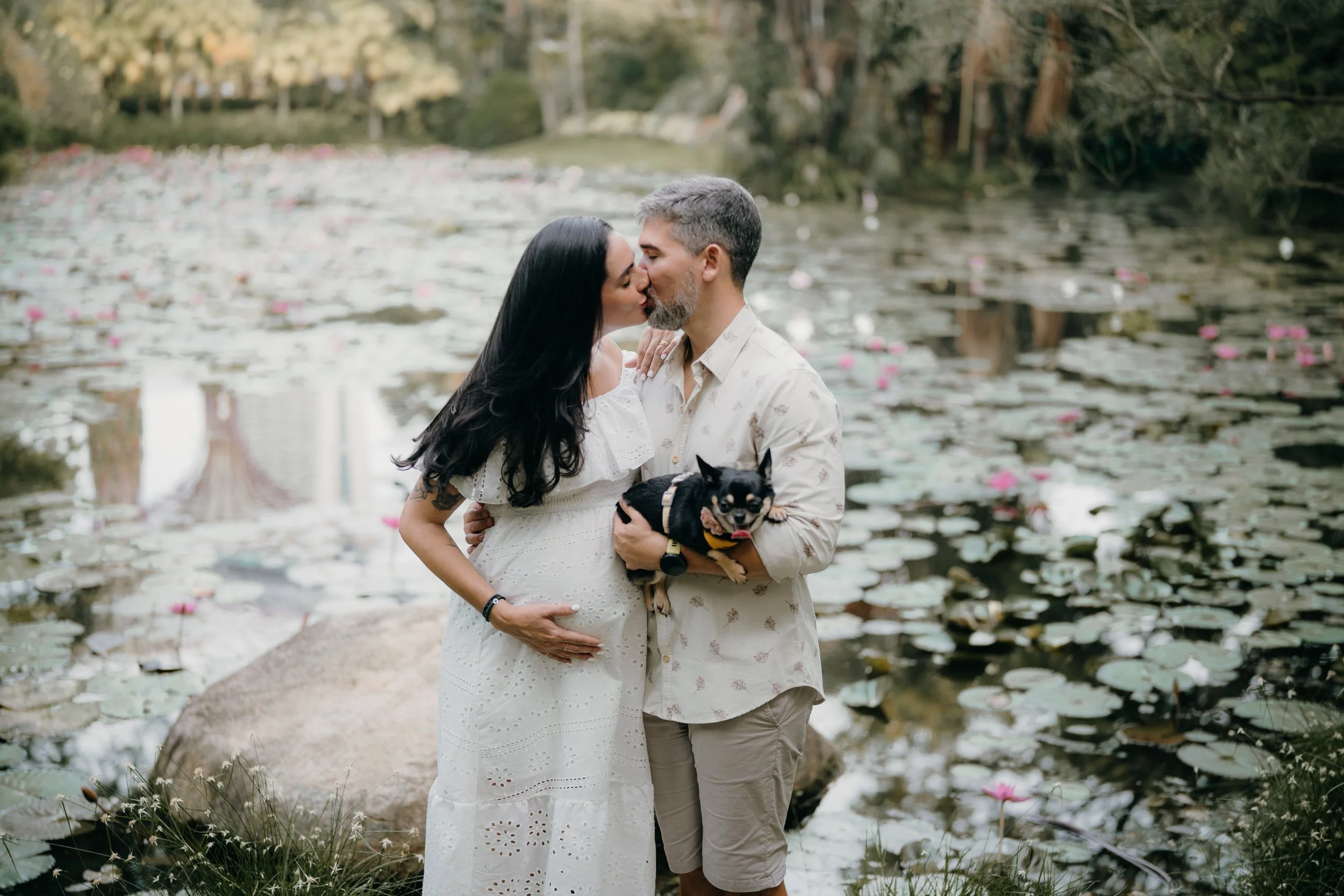 A couple kissing by a pond with water lilies, the woman is pregnant and holding her belly while the man holds a small dog.