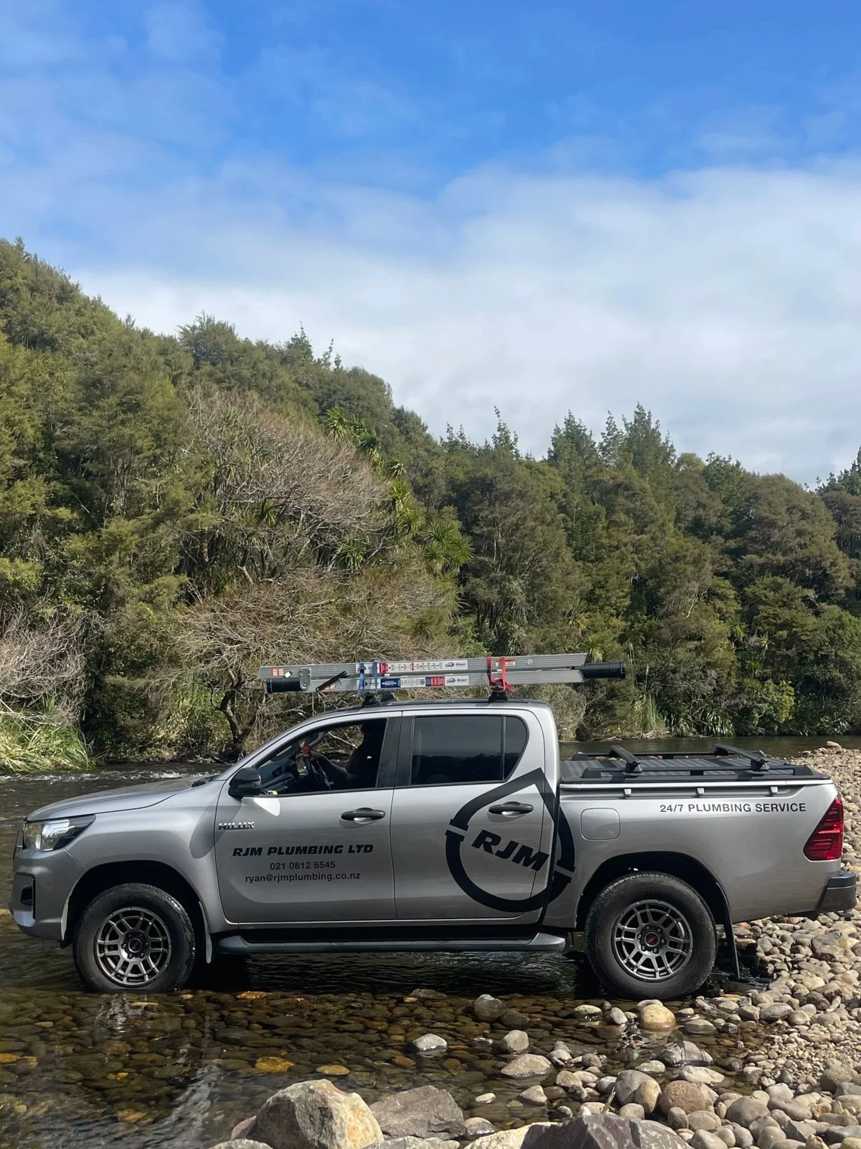 A silver pickup truck with 'RJM Plumbing Ltd' and contact information on the door parked in a shallow river with rocks, surrounded by green trees and hills under a partly cloudy sky.