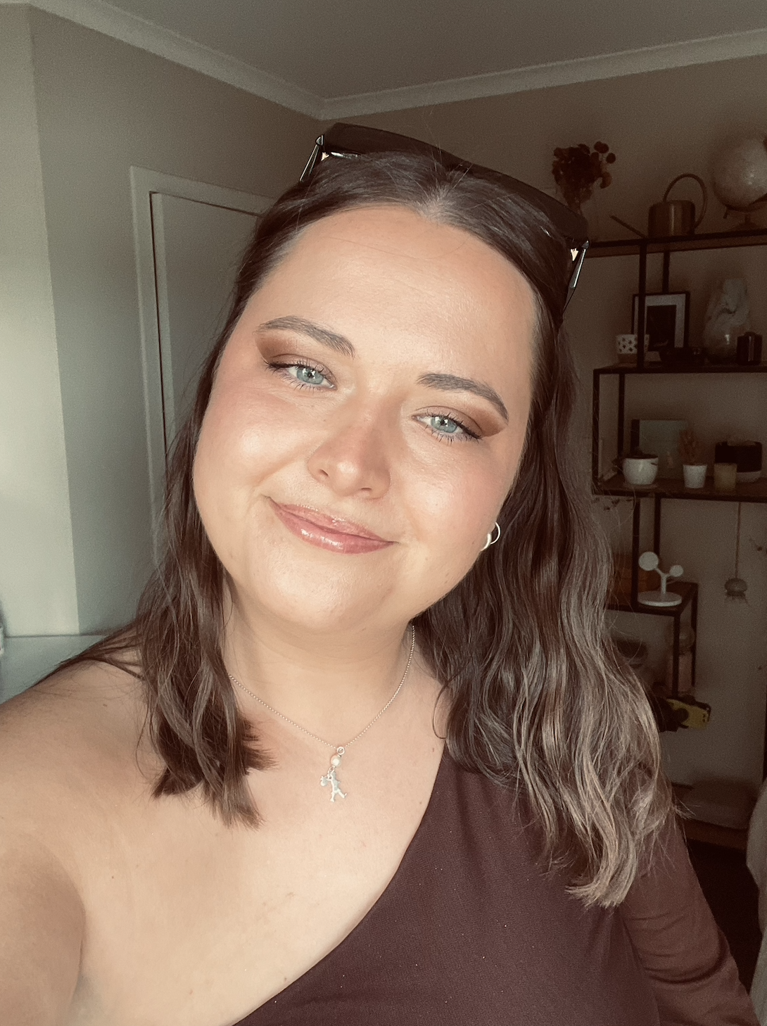 A woman with light skin and blue eyes taking a selfie indoors, wearing a dark top, a silver necklace, and glasses on her head, with shelves and decorative items in the background.