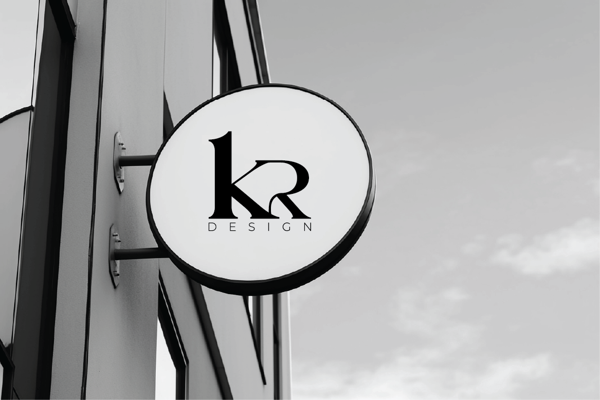 A circular sign on a building exterior displaying 'kr DESIGN' in black text on a white background, with a partly cloudy sky in the background.