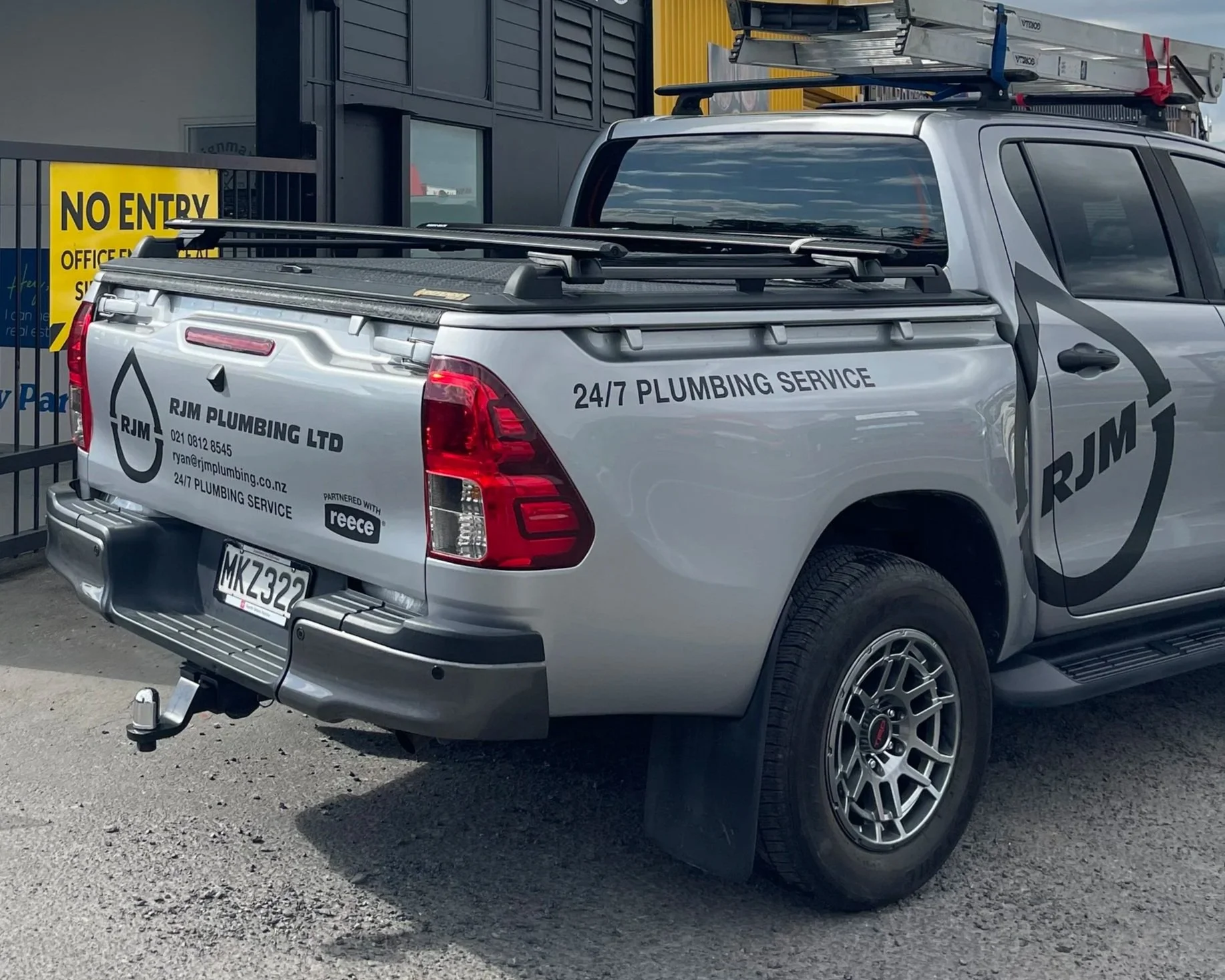 Silver pickup truck with RJM Plumbing Ltd branding, a ladder on top, parked behind a fence with a yellow NO ENTRY sign. The truck displays contact info and mentions 24/7 plumbing service.