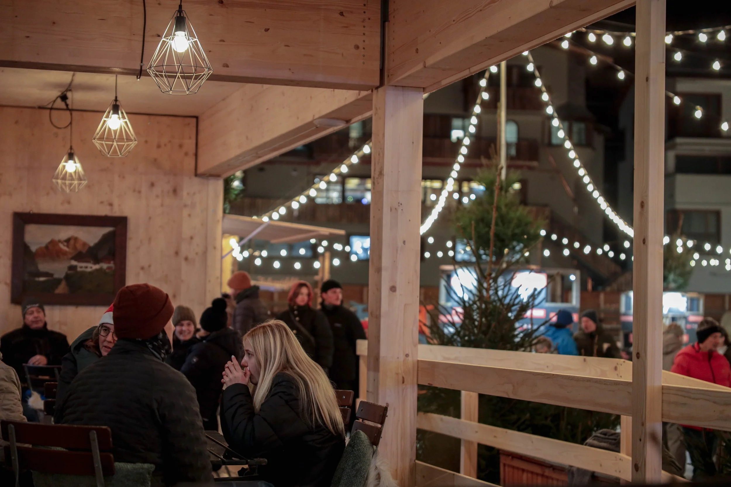 Menschen sitzen in einer festlich dekorierten, holzverkleideten Holzhütte mit Weihnachtsbaum, Lichterketten und Weihnachtsdekoration, bei Freiluftbetrieb am Abend.