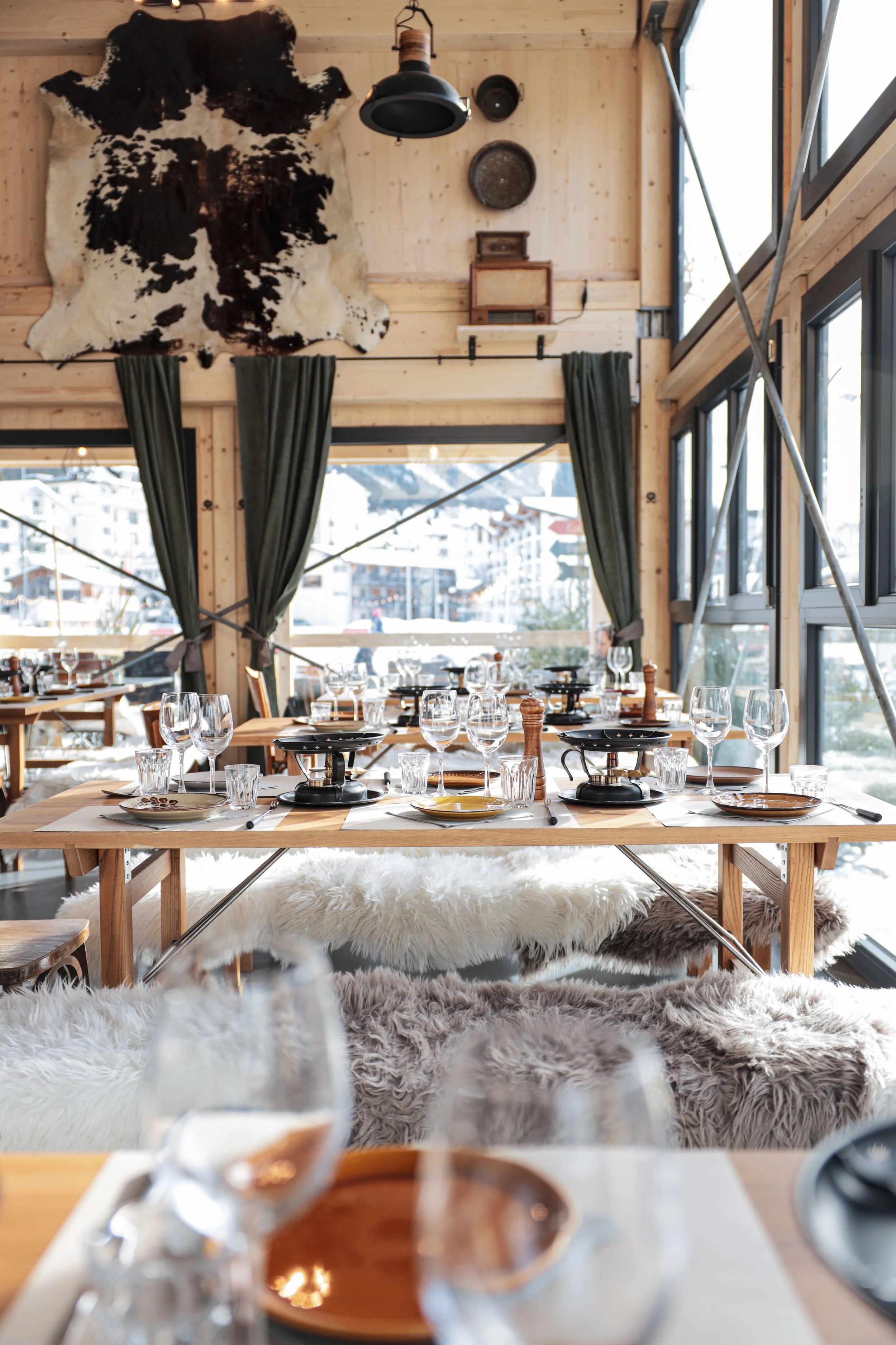 Ein gemütlich gedeckter Esstisch in einem Chalet mit Holzwänden, Felldecken, Gläsern, Tellern und einem Herd auf dem Tisch, mit großen Fenstern, die einen verschneiten Bergblick zeigen, und Wanddekorationen wie eine Kuhhaut und alte Gegenstände.