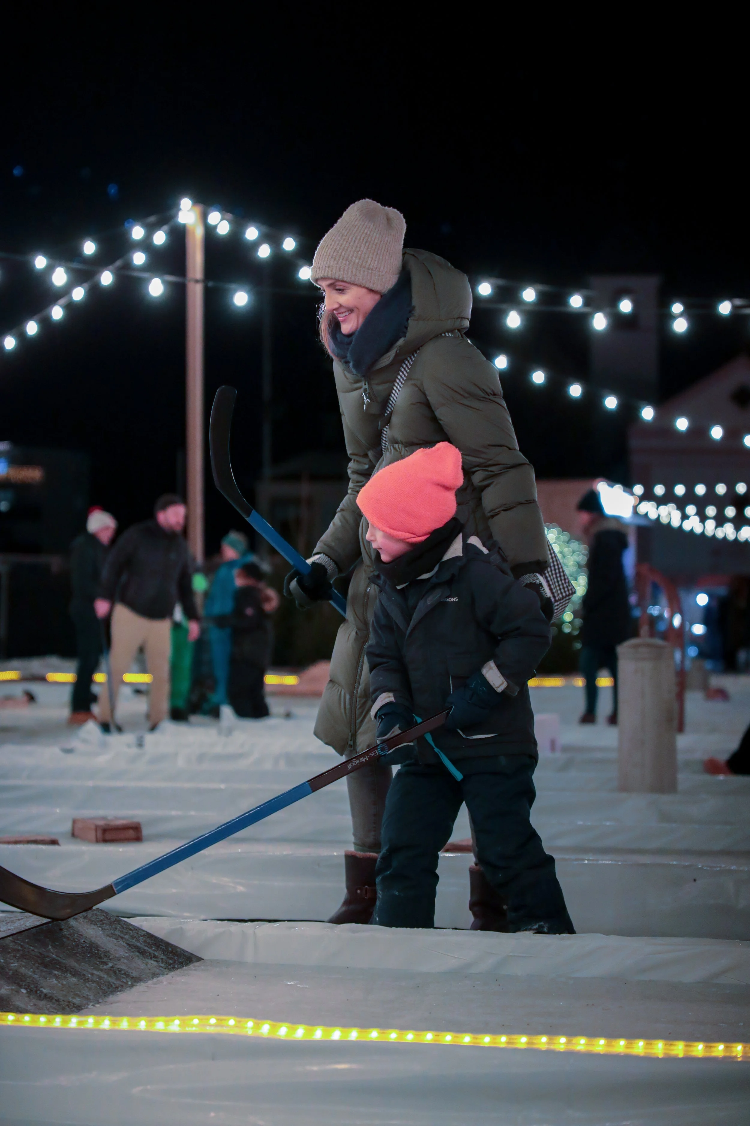 Eine Frau und ein Kind spielen Eishockey im Freien bei Nacht, beleuchtet von Lichterketten, im Winter.