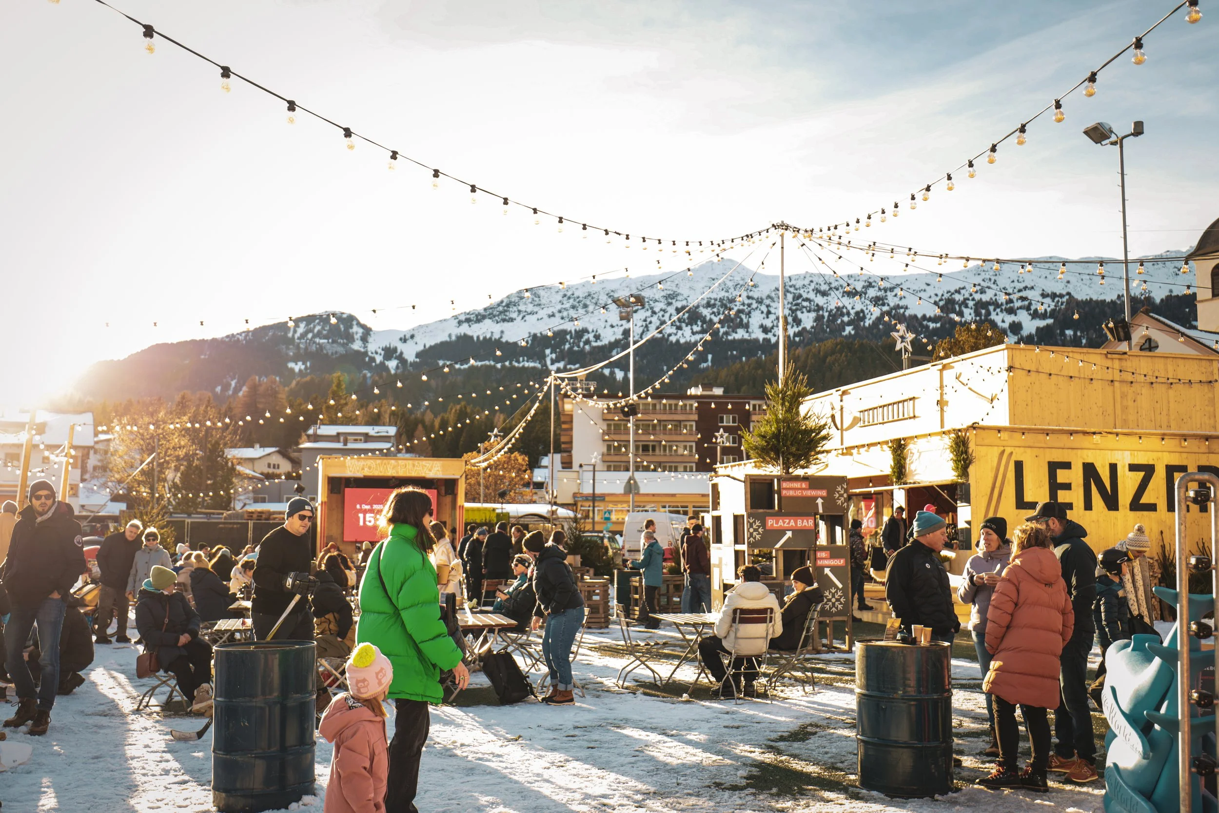 Menschen versammelt sich an einem winterlichen Outdoor-Markt oder Festival mit Schneebedeckten Bergen im Hintergrund, Lichterketten hängen über dem Platz, und die Leute tragen Winterkleidung wie Mäntel und Mützen.