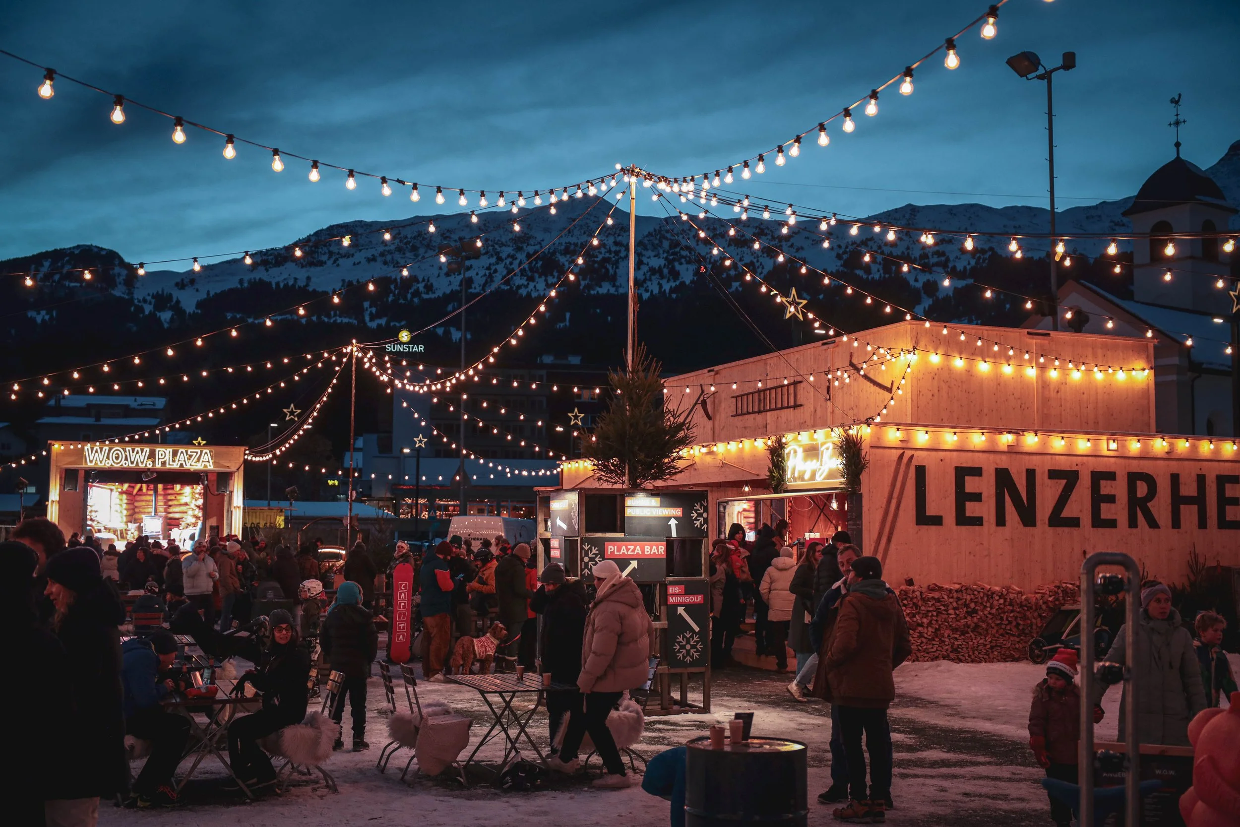 Weihnachtsmarkt in den Bergen bei Nacht mit Lichterketten, Menschen in dicken Jacken, Schnee und festlicher Beleuchtung.