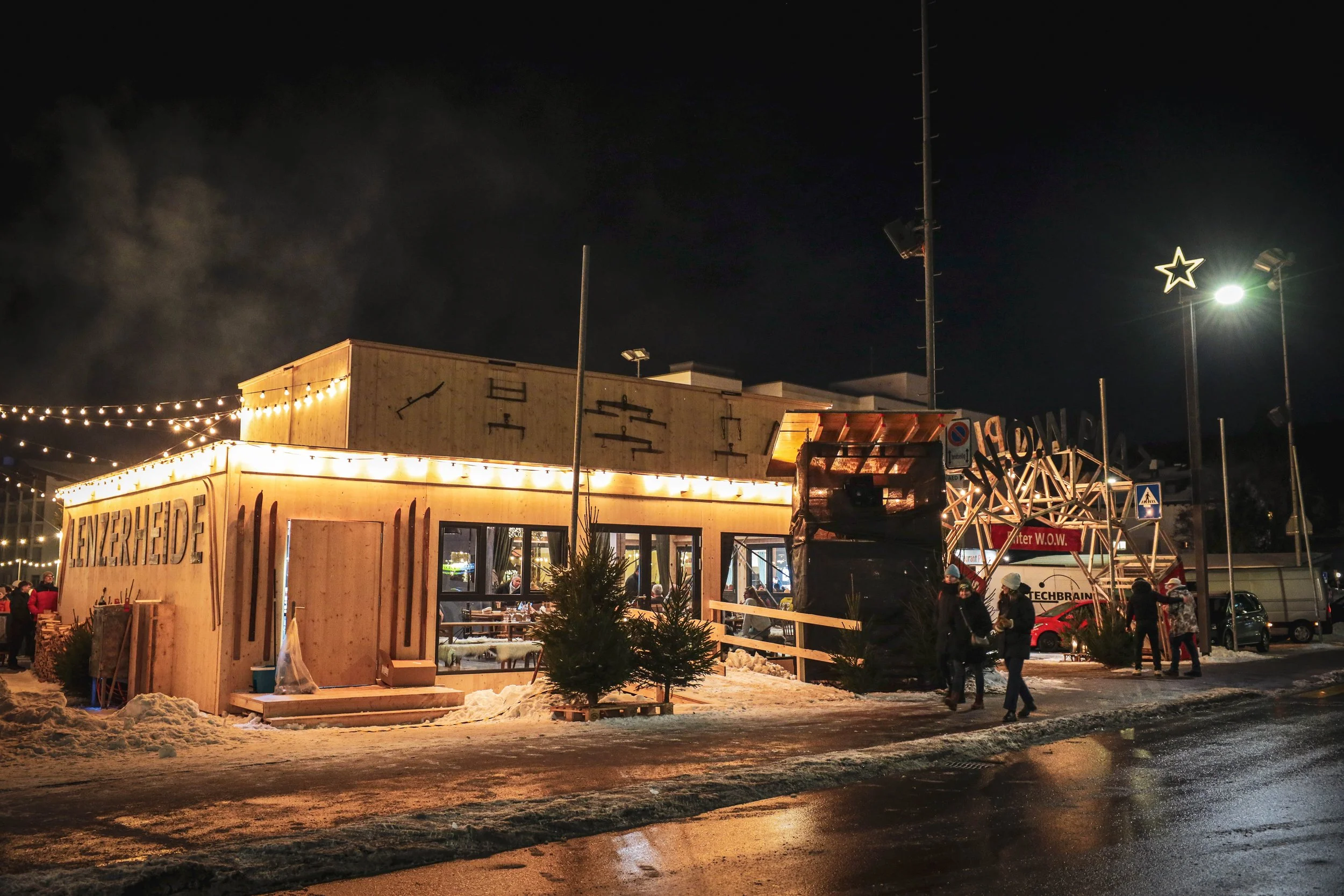 Ein beleuchteter Weihnachtsmarktstand im Winter bei Nacht, mit Schneedecke auf dem Boden und mehreren Besuchern davor.
