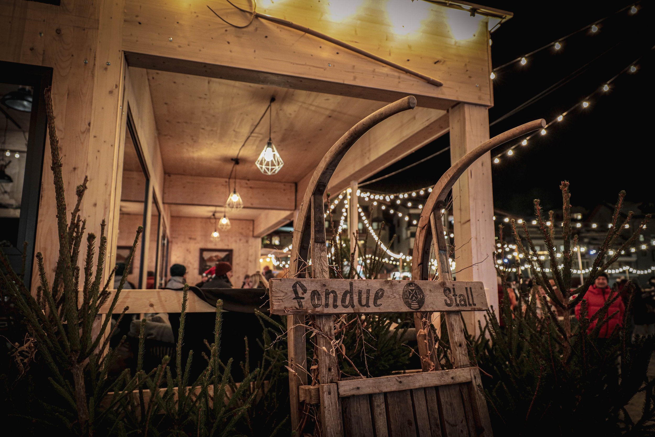 Holzstall mit Schild 'Fondue Stall' im festlichen Lichterglanz bei Nacht, umgeben von Tannenzweigen, in einer winterlichen Weihnachtsmarkt-Szene.