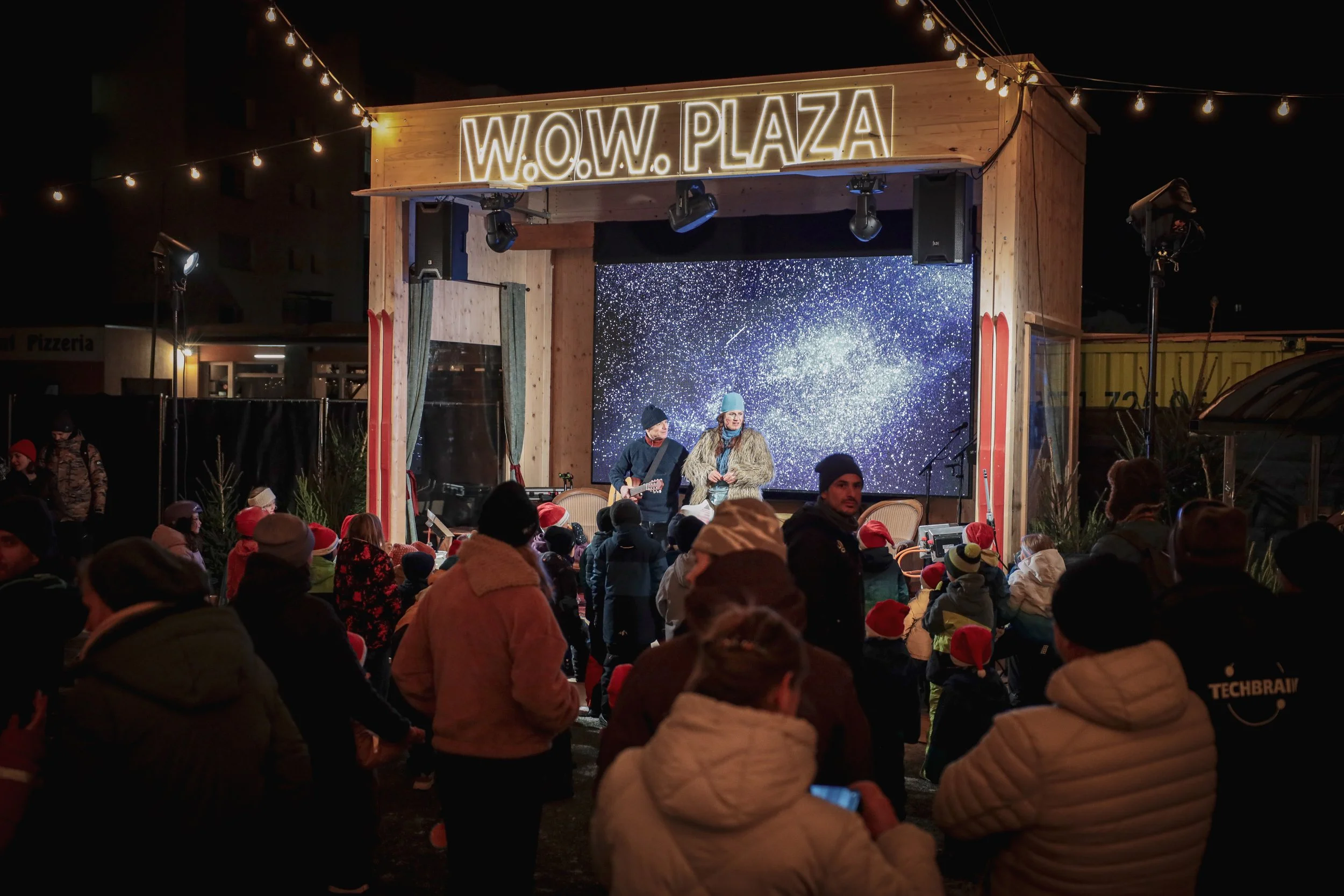 Eine festliche Weihnachtsveranstaltung im Freien mit einer Bühne, auf der eine Band auftritt. Das Publikum trägt Weihnachtsmützen, einige fotografieren oder filmen die Darbietung. Es sind Lichter und Dekorationen sichtbar, die eine festliche Atmosphä