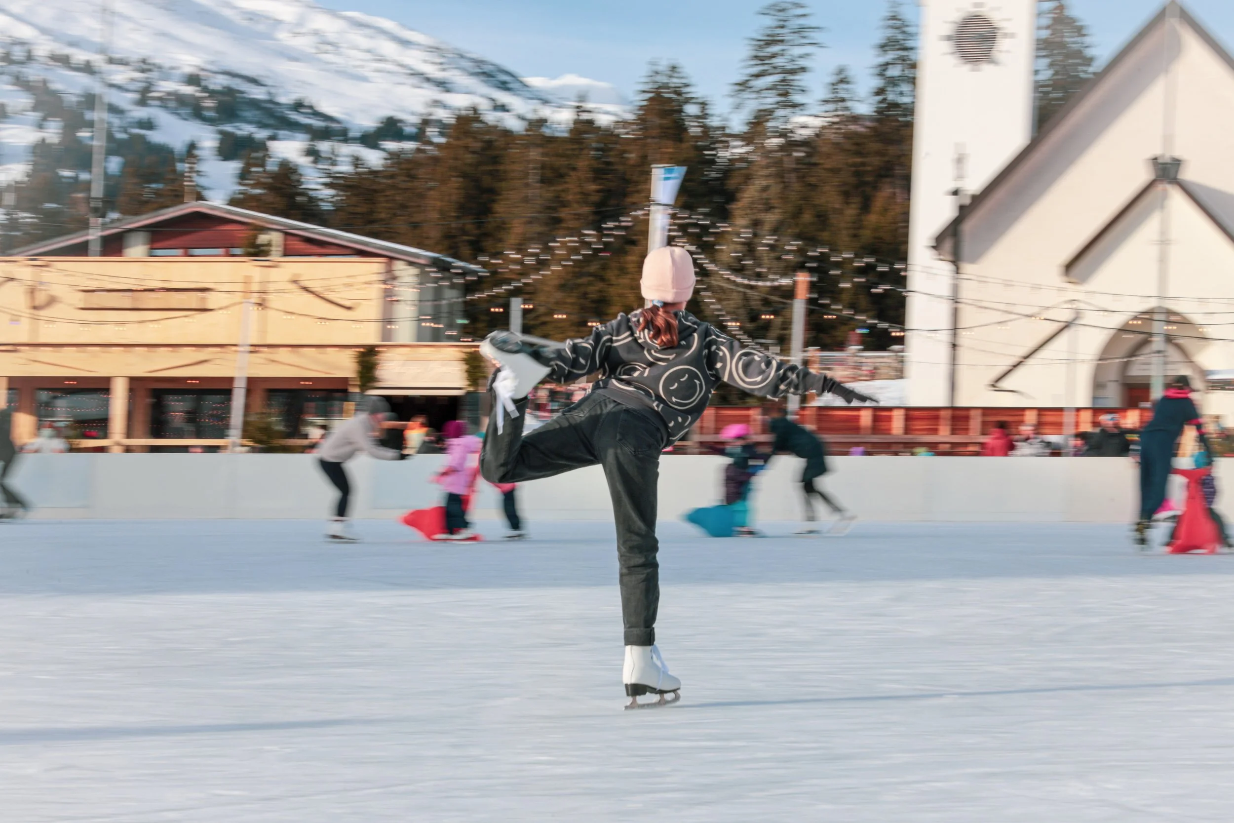 Junge beim Eislaufen auf einer Eisbahn, im Hintergrund sind andere Eisläufer und eine Kirche sowie Berge im Winter.