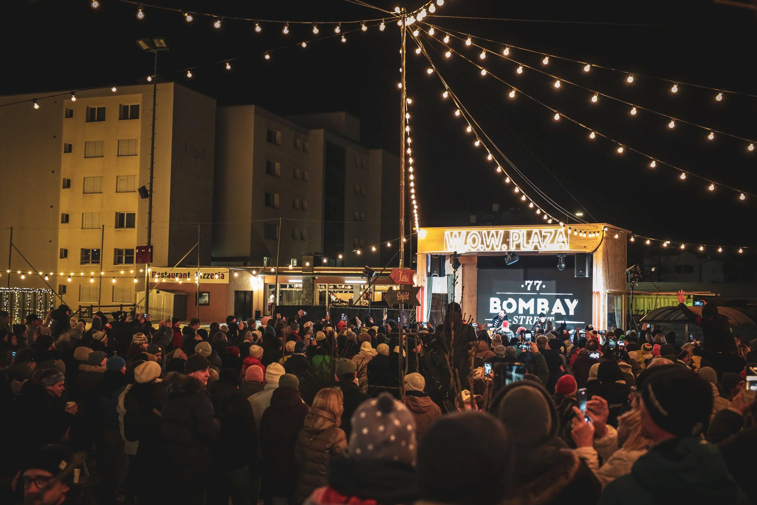 Menschen versammeln sich bei einem nächtlichen Open-Air-Konzert mit Lichterketten, ein großer Bildschirm zeigt 'BOMBAY' und eine Bühne mit Musikern.
