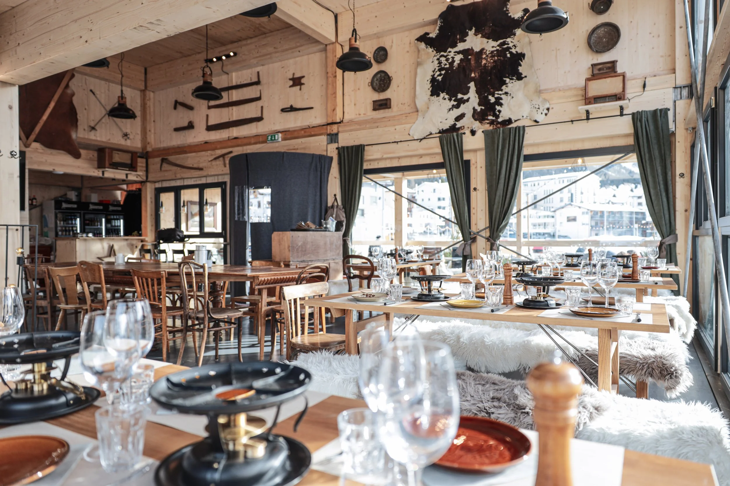 Inneneinrichtung eines rustikalen Restaurants mit Holzmöbeln, großen Fenstern, grünem Vorhang, Tierfellen und dekorativen Gegenständen an der Wand.