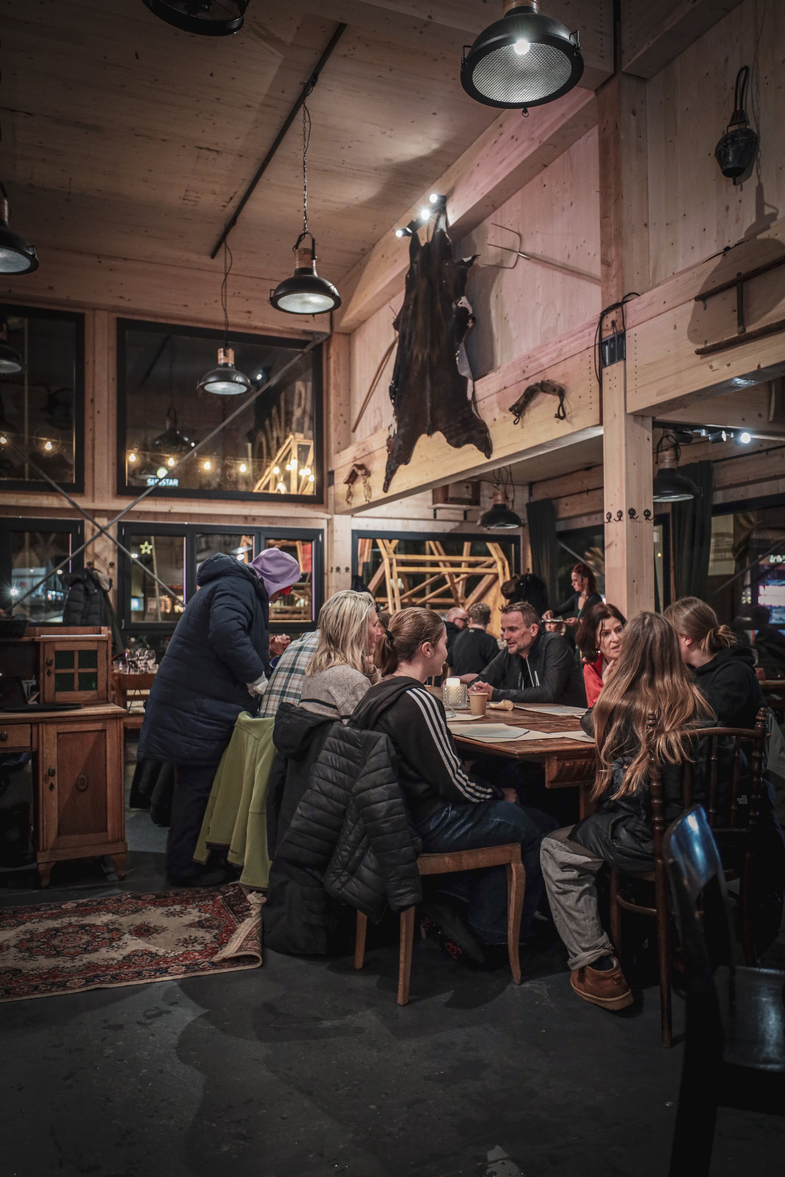 Menschen sitzen in einem rustikalen Restaurant mit Holzmöbeln und Tierfellen an der Wand, während im Hintergrund weitere Gäste speisen.