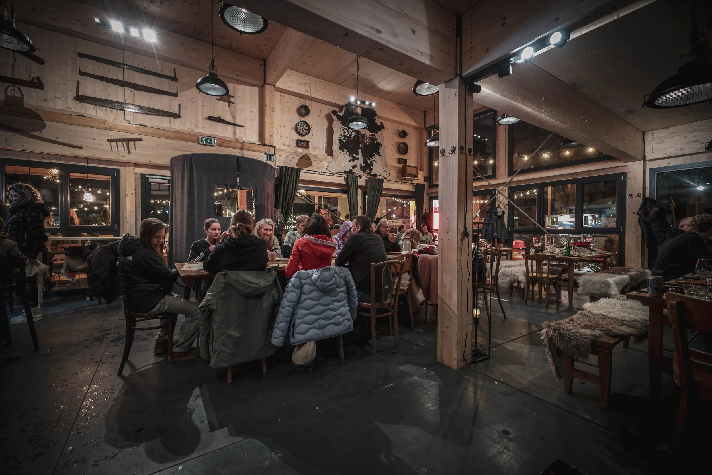 Innenaufnahme eines gemütlichen Restaurants mit mehreren Tischen, an denen Menschen sitzen und essen. Das Interieur besteht aus Holz mit rustikalem Dekor und warmem Licht.