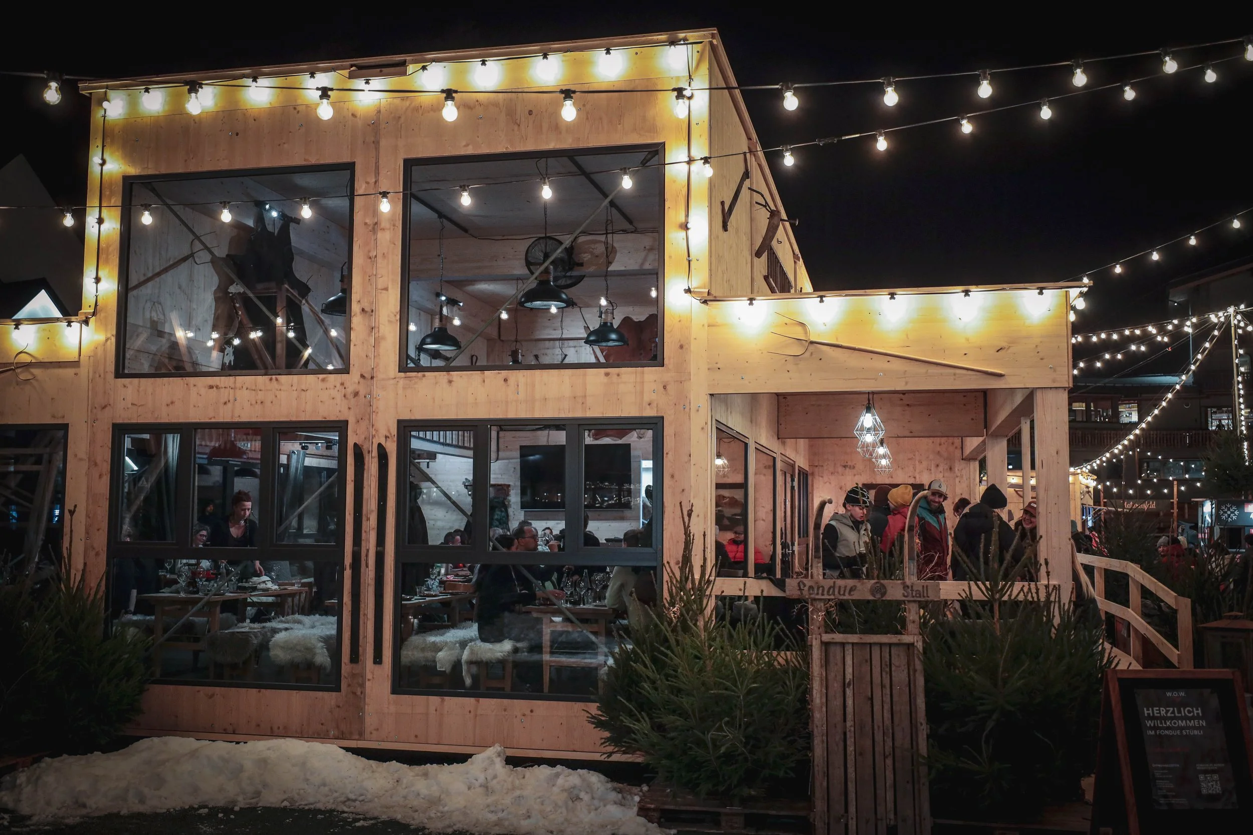 Ein Holzgebäude mit Fenstern, das bei Nacht mit Lichterketten dekoriert ist. Es gibt Menschen, die im Inneren und auf der Terrasse sitzen, und draußen stehen Leute.